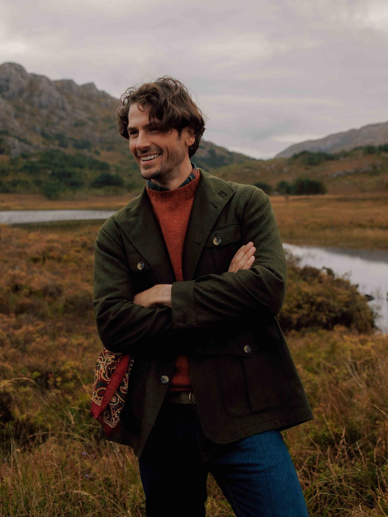 A man with wavy hair stands outdoors in a grassy setting, smiling with arms crossed. He wears the Campbells of Beauly Field Teba Jacket, a rust-red sweater, jeans, and carries a patterned bag over his shoulder.