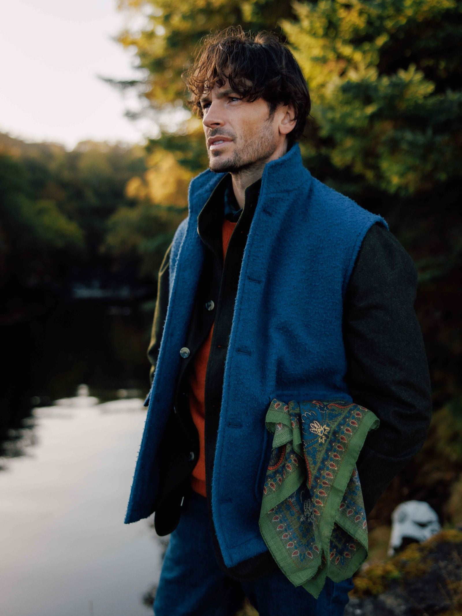 A man with wavy hair stands by a calm lake, wearing the Campbell's of Beauly Casentino Fleece Gilet over a dark shirt and orange sweater, holding a green patterned scarf amid trees in a natural outdoor setting.