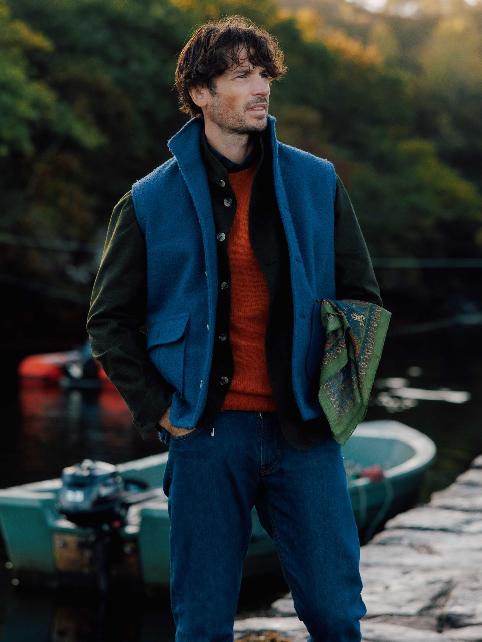 A man stands by a docked boat and water, wearing the Campbell's of Beauly Casentino Fleece Gilet over a red sweater and dark shirt with blue jeans. He holds a green patterned scarf amid trees and soft sunlight, looking to the side.