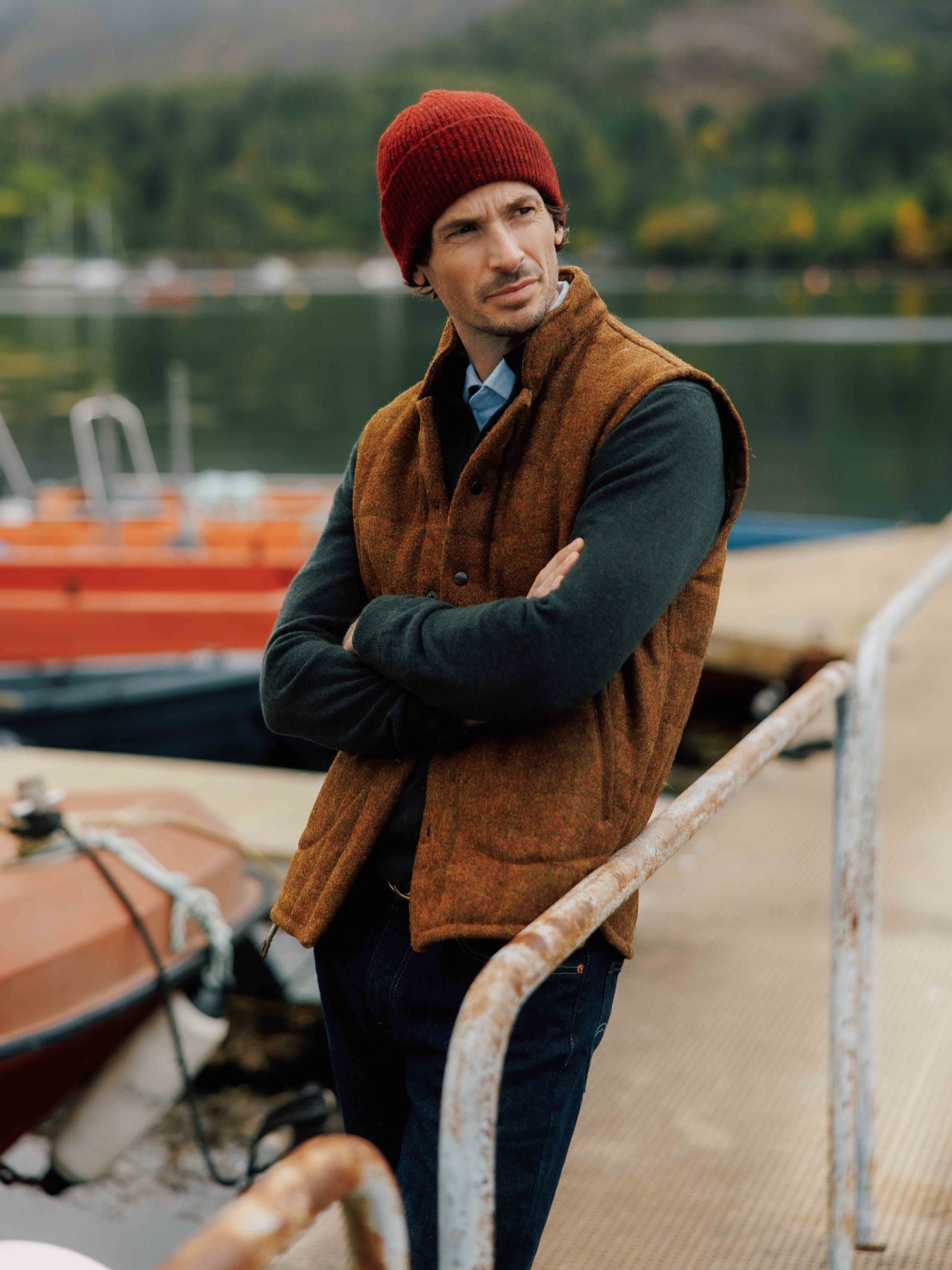 A man in a Campbell's of Beauly Harris Tweed Quilted Gilet, dark sweater, and red beanie stands on a dock beside orange boats, with calm lake waters and forested hills behind him.