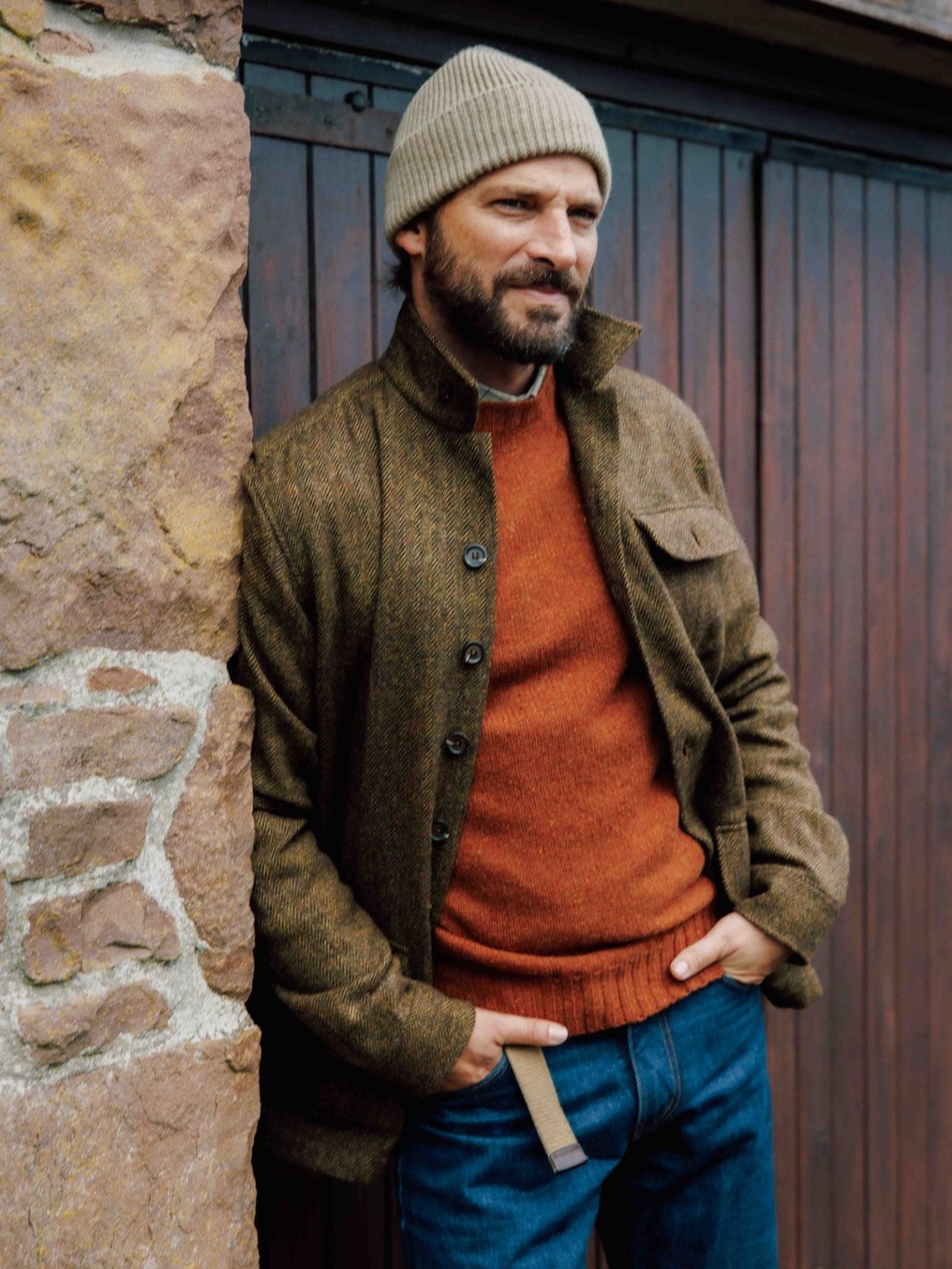 A bearded man in a beige knit beanie, brown jacket, blue jeans, and a Campbell's of Beauly Donegal Lambswool Crew Neck Jumper stands by a stone wall with hands in pockets, looking to the side.