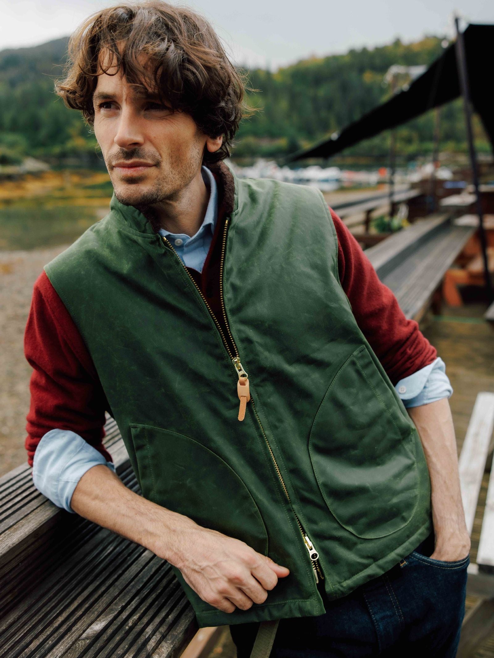 A man with wavy brown hair wears the Campbell's of Beauly Wax Nehru Gilet, a green waxed cotton zip-up vest, over a red sweater and blue shirt while leaning against a wooden railing in a scenic countryside setting with trees and water.