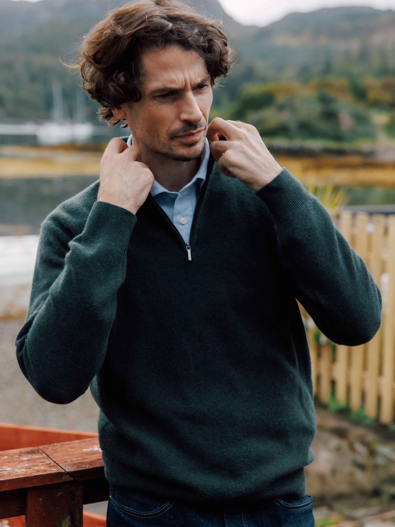 A man with curly brown hair stands outdoors, looking thoughtful as he adjusts the collar of his Campbell's of Beauly Piquet Quarter Zip Jumper in dark green, layered over a light blue shirt. A wooden fence and trees blur in the background.