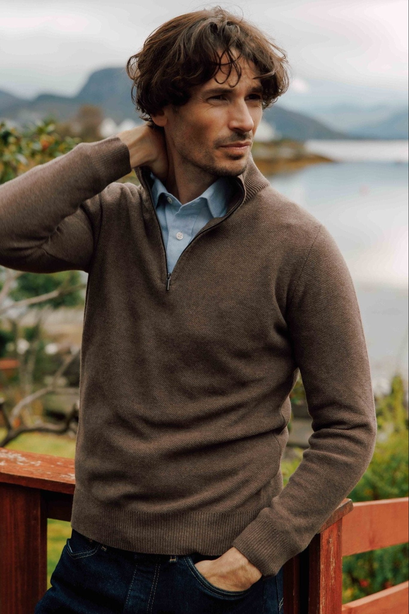 A man with wavy brown hair stands outdoors by a wooden railing, wearing a Campbell's of Beauly Auld Stock Piquet Quarter Zip Jumper over a light blue shirt, looking to the side with one hand behind his neck. A lake and mountains are in the background.