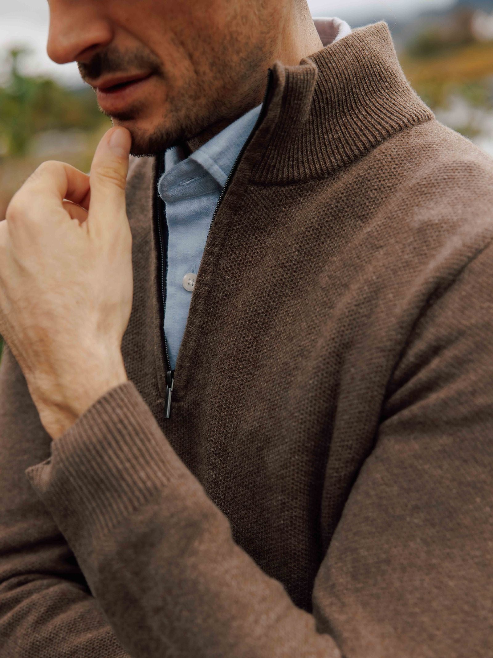 A man wears the Campbell's of Beauly Piquet Quarter Zip Jumper over a light blue shirt, standing outdoors with his hand on his chin. His partially visible face and a blurred background enhance the contemplative atmosphere.