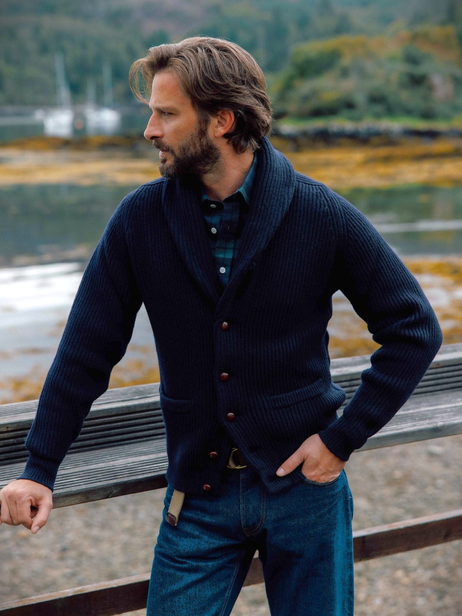A man with light brown hair and a beard stands outdoors by a wooden railing, wearing the Campbell's of Beauly Shawl Collar Cardigan in dark blue over a plaid shirt and jeans, with scenic water and forest in the background.