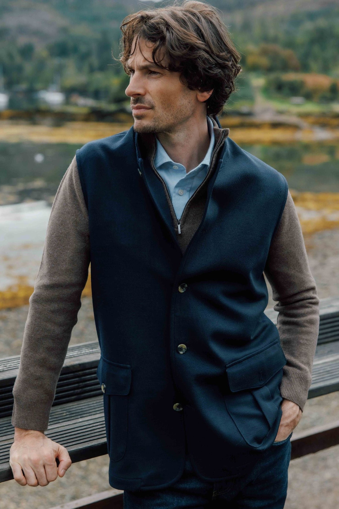 A man with wavy brown hair stands outdoors by water, wearing the Campbell's of Beauly Knitted Nehru Gilet over a brown sweater and light blue collared shirt, looking to the side with one hand in his pocket; trees and hills are behind.
