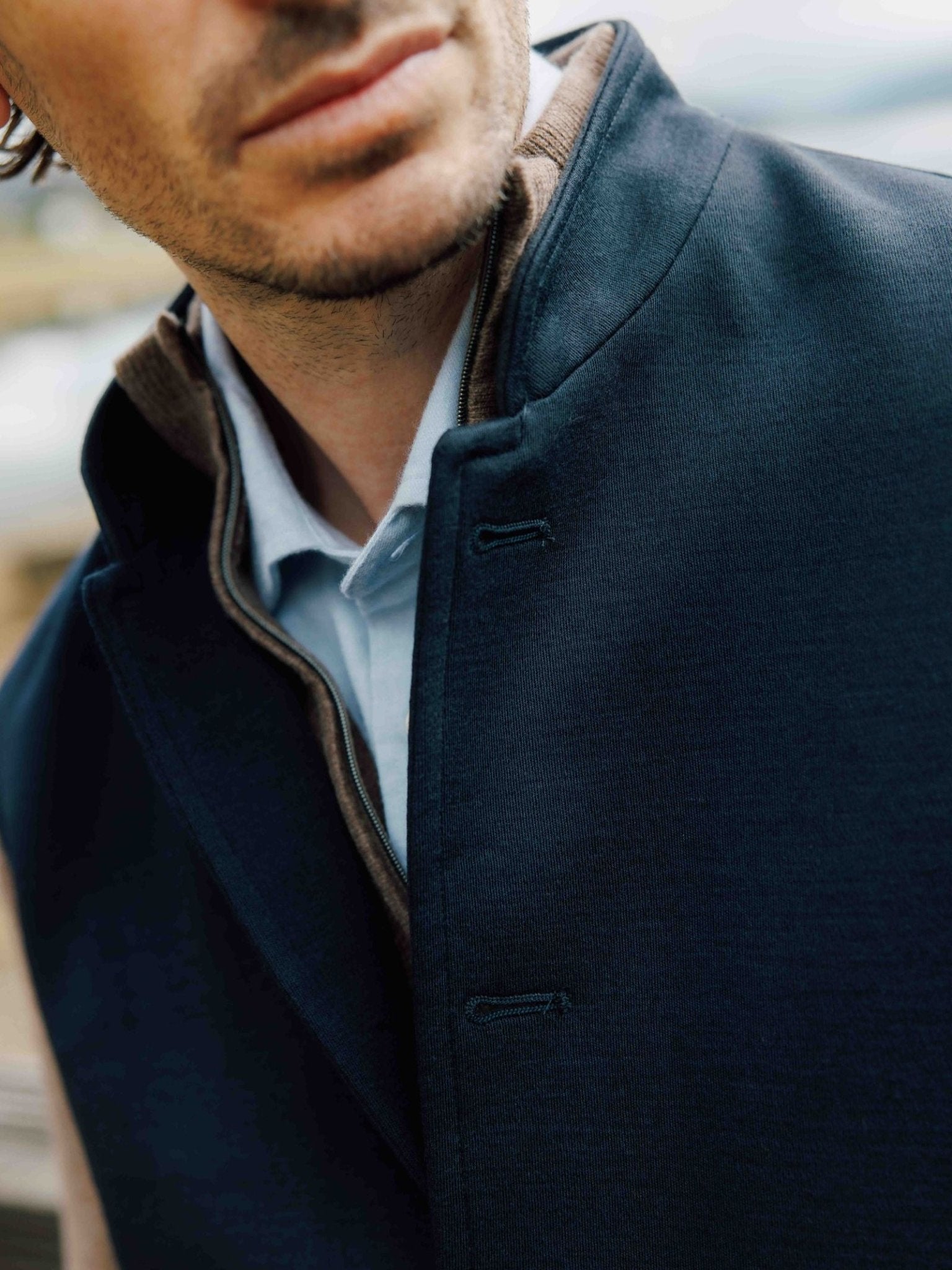 Close-up of a man in a dark blue coat over a light blue shirt and Campbell’s of Beauly Auld Stock Knitted Nehru Gilet, crafted from 100% wool. His face is partially visible with a light beard; the blurred background highlights its heritage style.