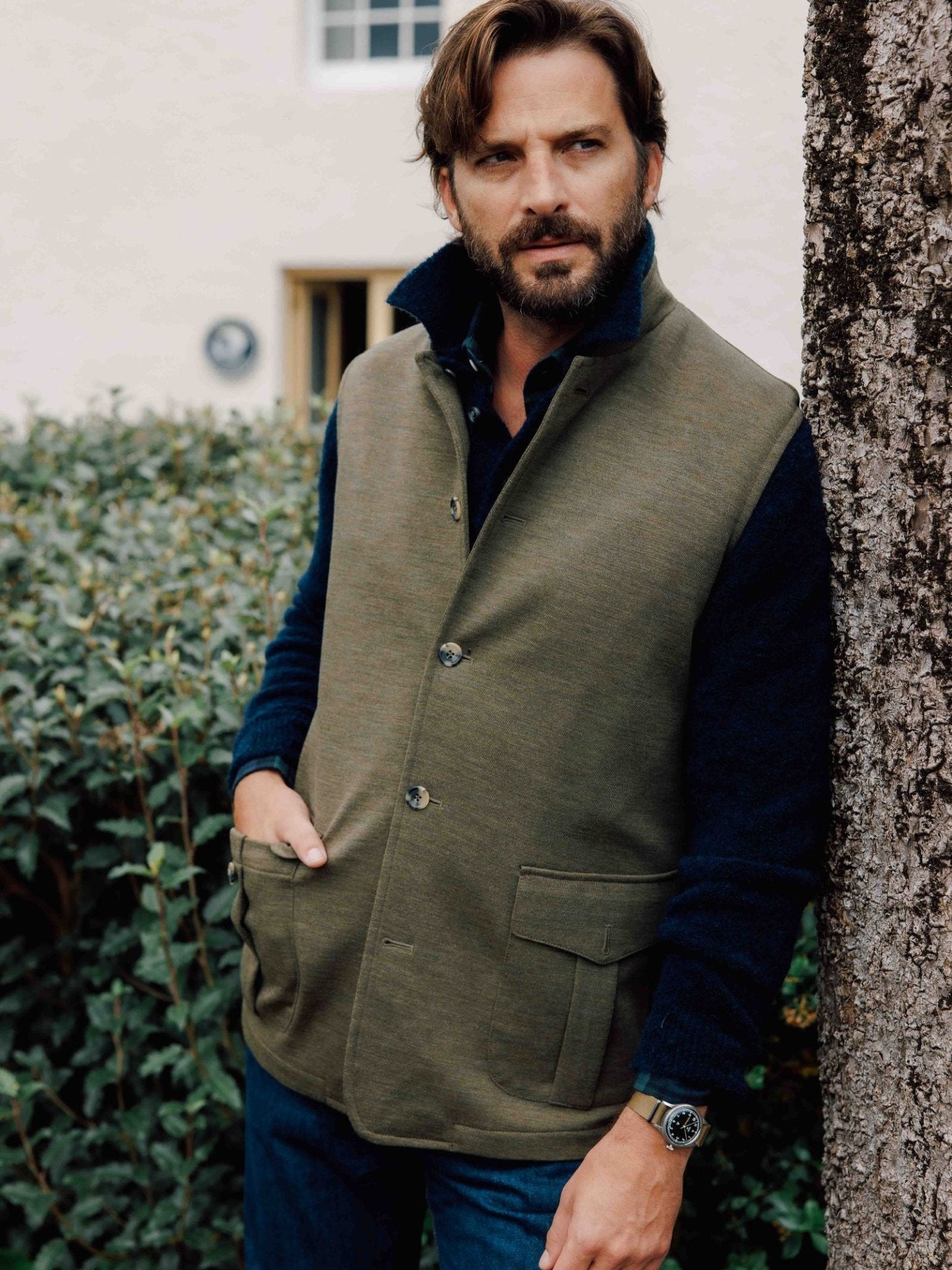A man with brown hair and a beard stands outdoors by a tree, wearing the Campbell's of Beauly Knitted Nehru Gilet in green over a dark shirt and blue jeans, his left hand in his pocket, with shrubs and a white building behind him.