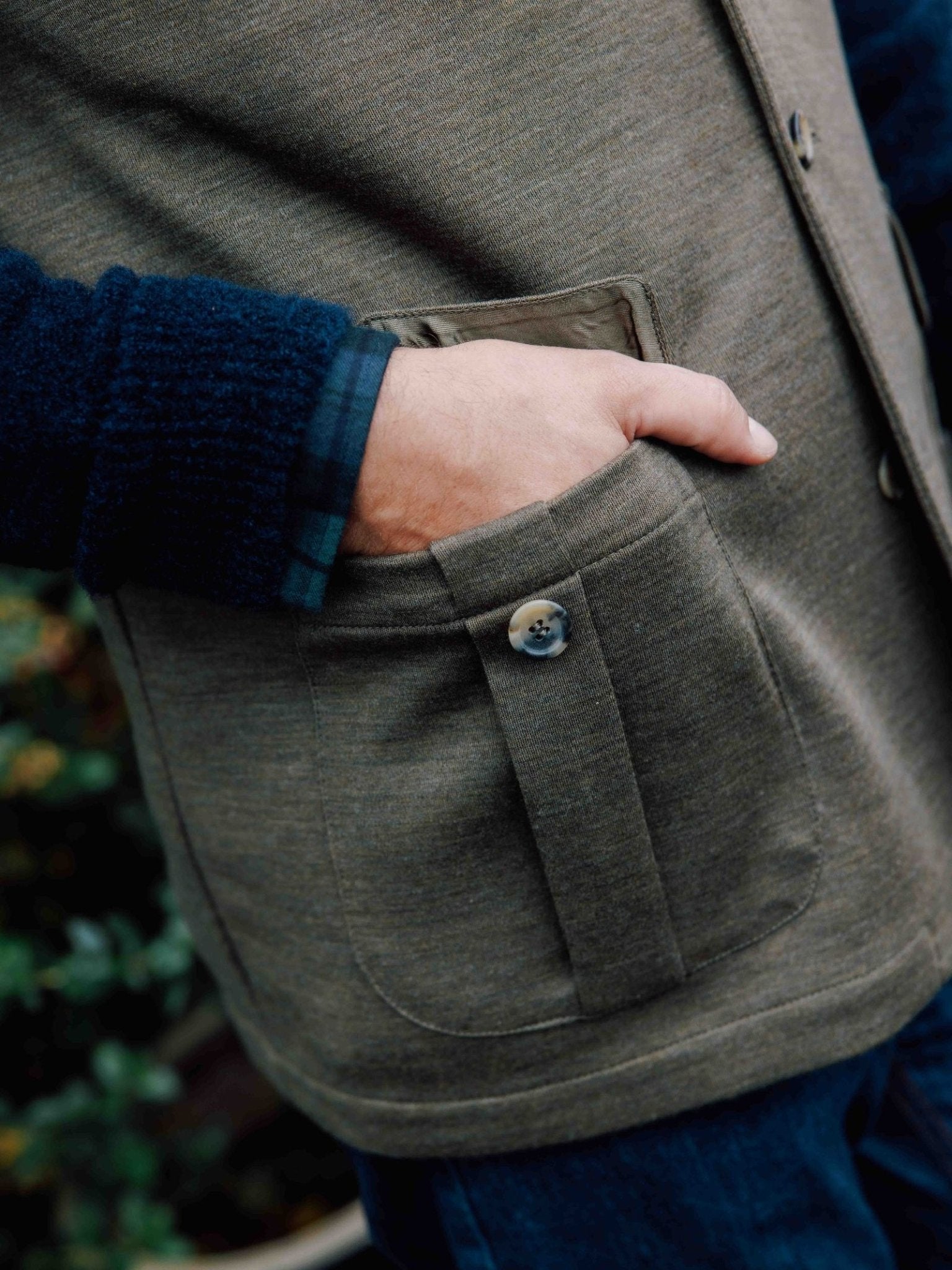 A person wearing the Campbell's of Beauly Auld Stock Knitted Nehru Gilet in a heritage greenish-brown, with a blue plaid shirt underneath, stands outdoors with one hand in the buttoned front pocket. Only their torso and arm are visible.