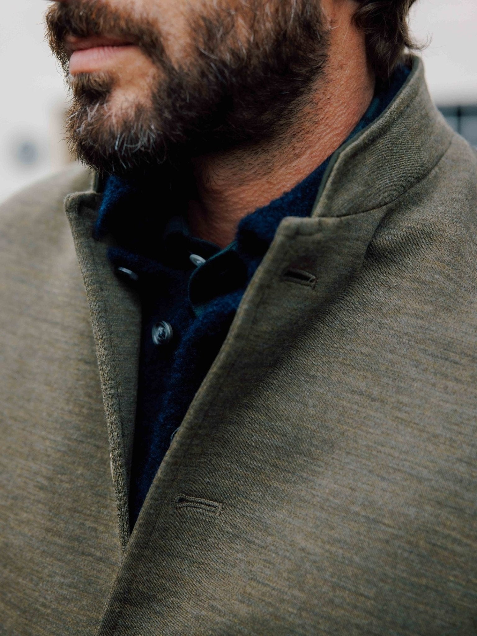 Close-up of a man's bearded lower face wearing the Campbell's of Beauly Auld Stock Knitted Nehru Gilet in olive green wool over a dark shirt, with focus on the textured fabric and collar detail, evoking classic heritage style against a blurred backdrop.