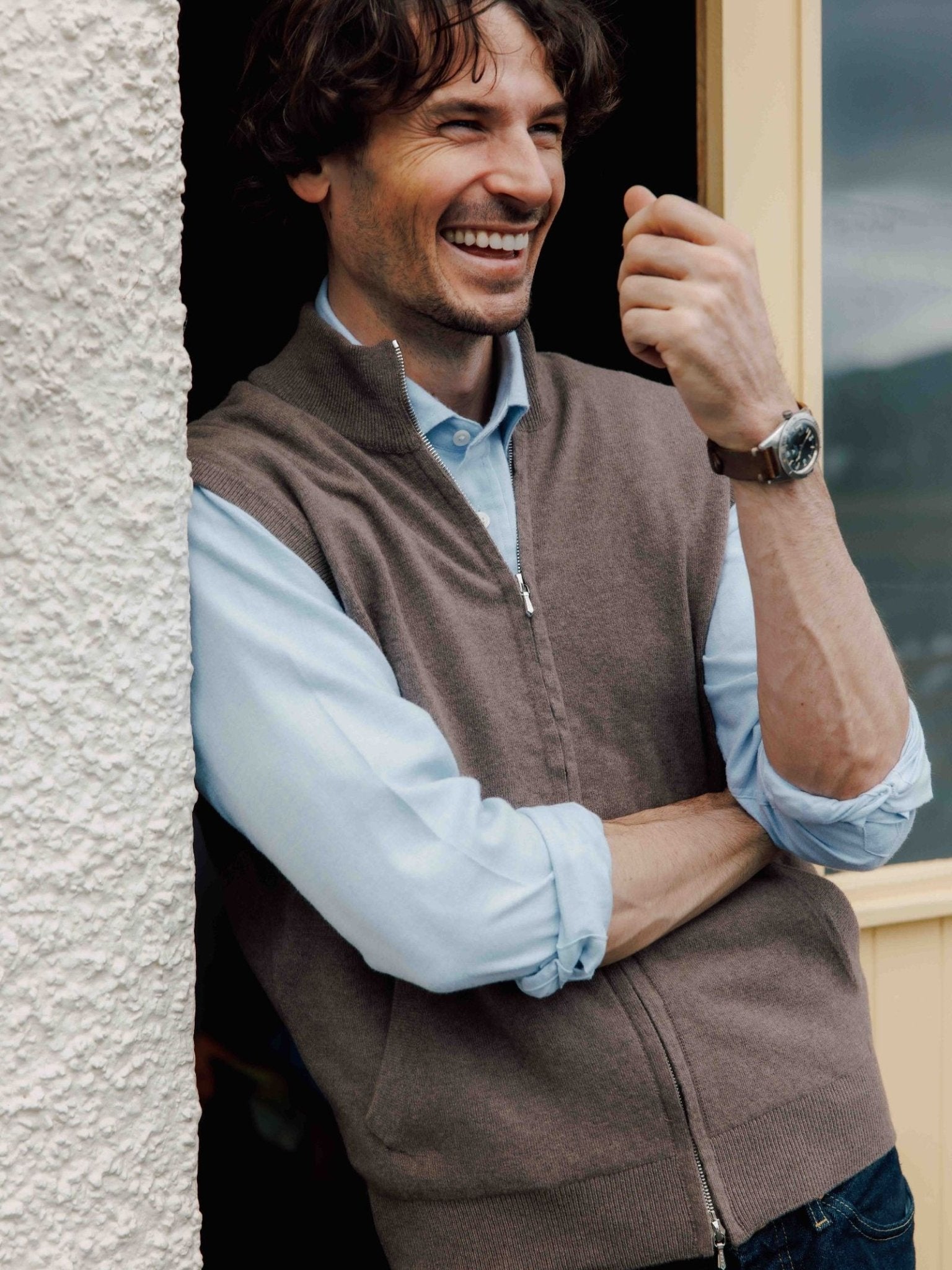A smiling man with wavy hair leans against a textured wall, wearing the Twill Shirt by Campbell's of Beauly in light blue and a brown zip-up vest. He crosses his arms and raises one hand to showcase a wristwatch.