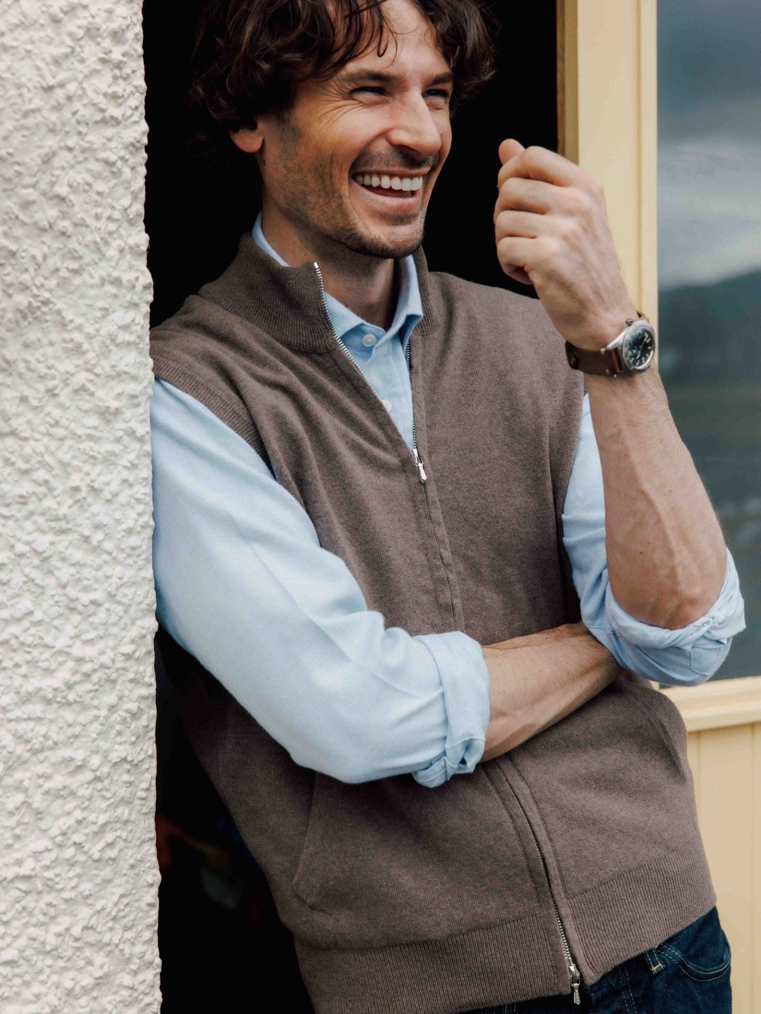 A man with wavy brown hair, wearing a light blue shirt and a Campbell's of Beauly Zip Gilet, stands by a textured wall, smiling with his arms crossed and one hand raised.