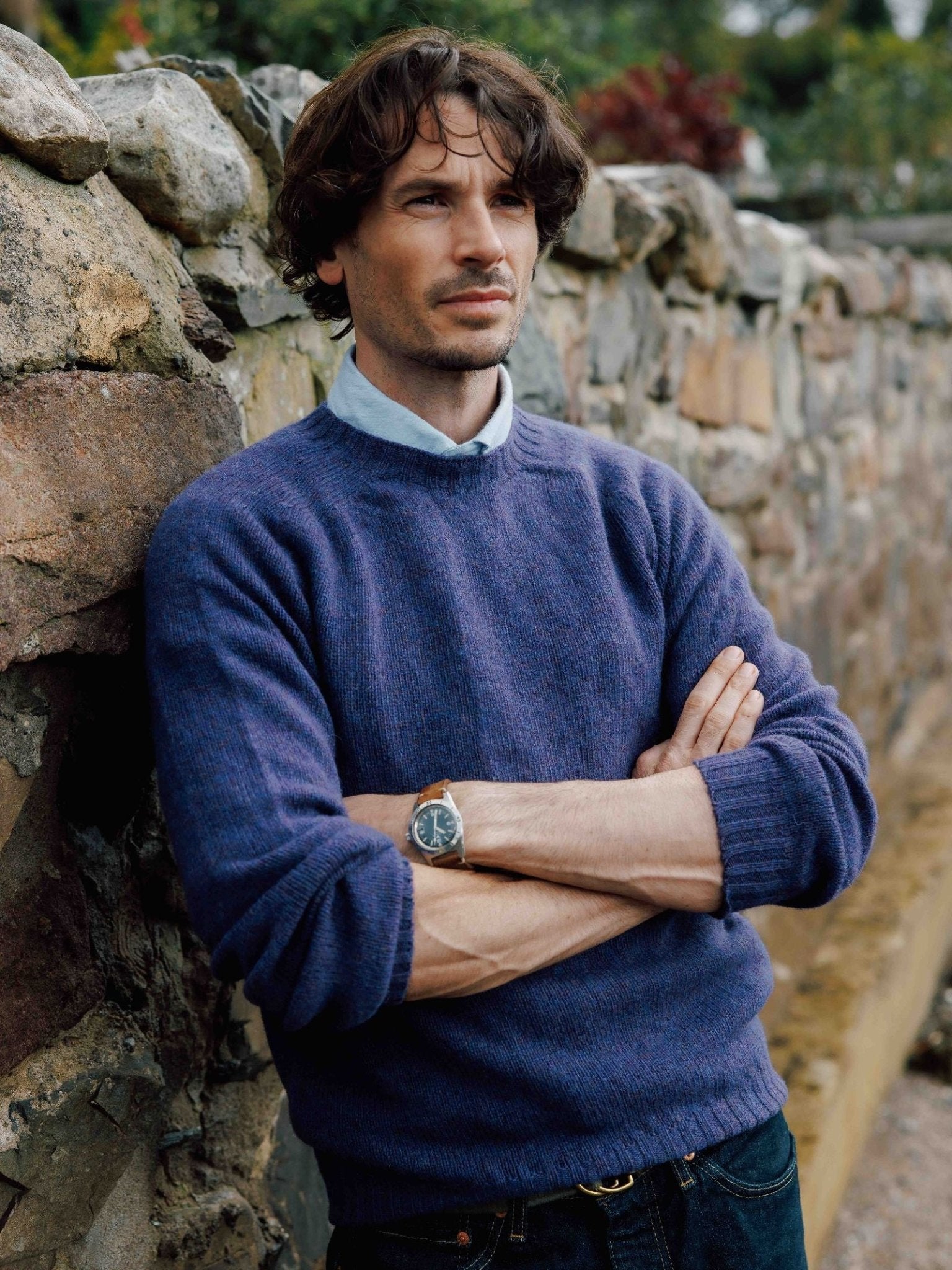 A man with wavy brown hair, wearing a Campbell's of Beauly Shetland Jumper in blue over a collared shirt, stands with arms crossed against a rustic stone wall outdoors, looking ahead with a serious expression.