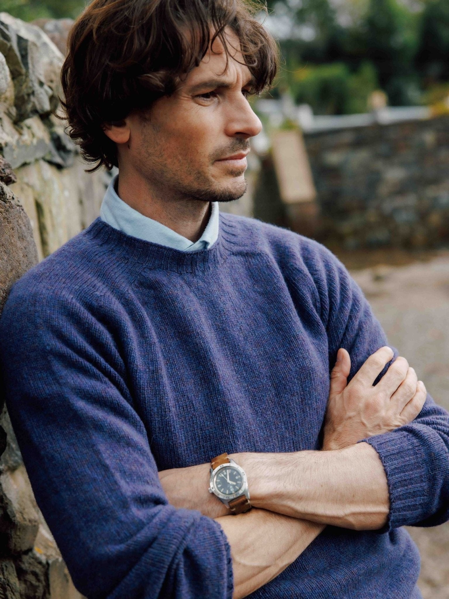 A man with wavy brown hair stands outdoors, arms crossed and leaning against a stone wall. He wears a light blue shirt paired with the Campbell's of Beauly Shetland Jumper and a wristwatch, gazing thoughtfully into the distance.