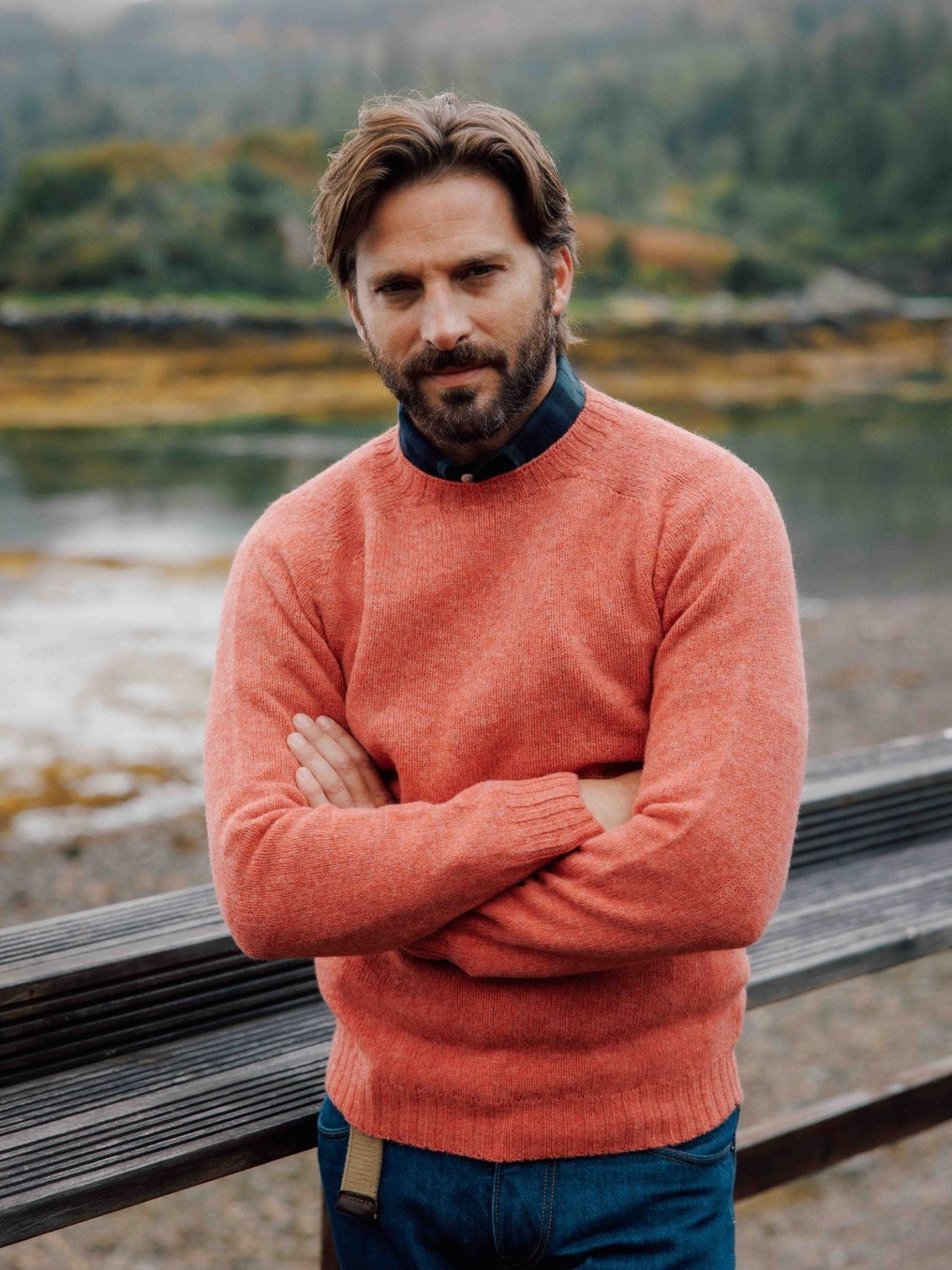 A dark-haired, bearded man stands outdoors with arms crossed, wearing a coral Campbell's of Beauly Shetland Jumper over a dark shirt, in front of a wooden railing with water, trees, and hills softly blurred in the background.