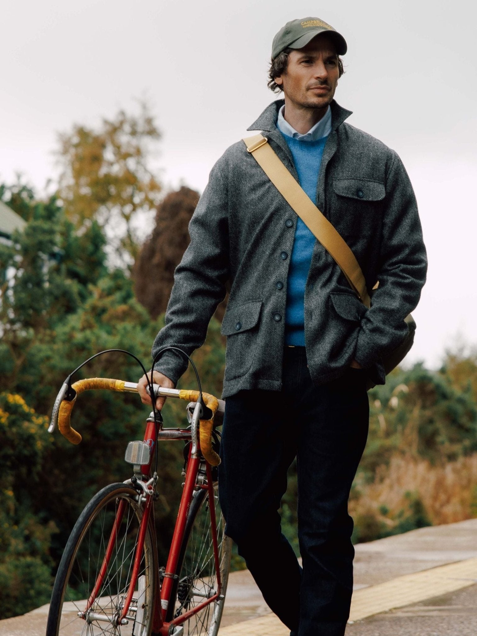 A man in a gray Casual Shacket by Campbell's of Beauly, blue sweater, dark jeans, and green cap walks outdoors with a red bicycle and yellow strap bag over his shoulder, greenery in the background.