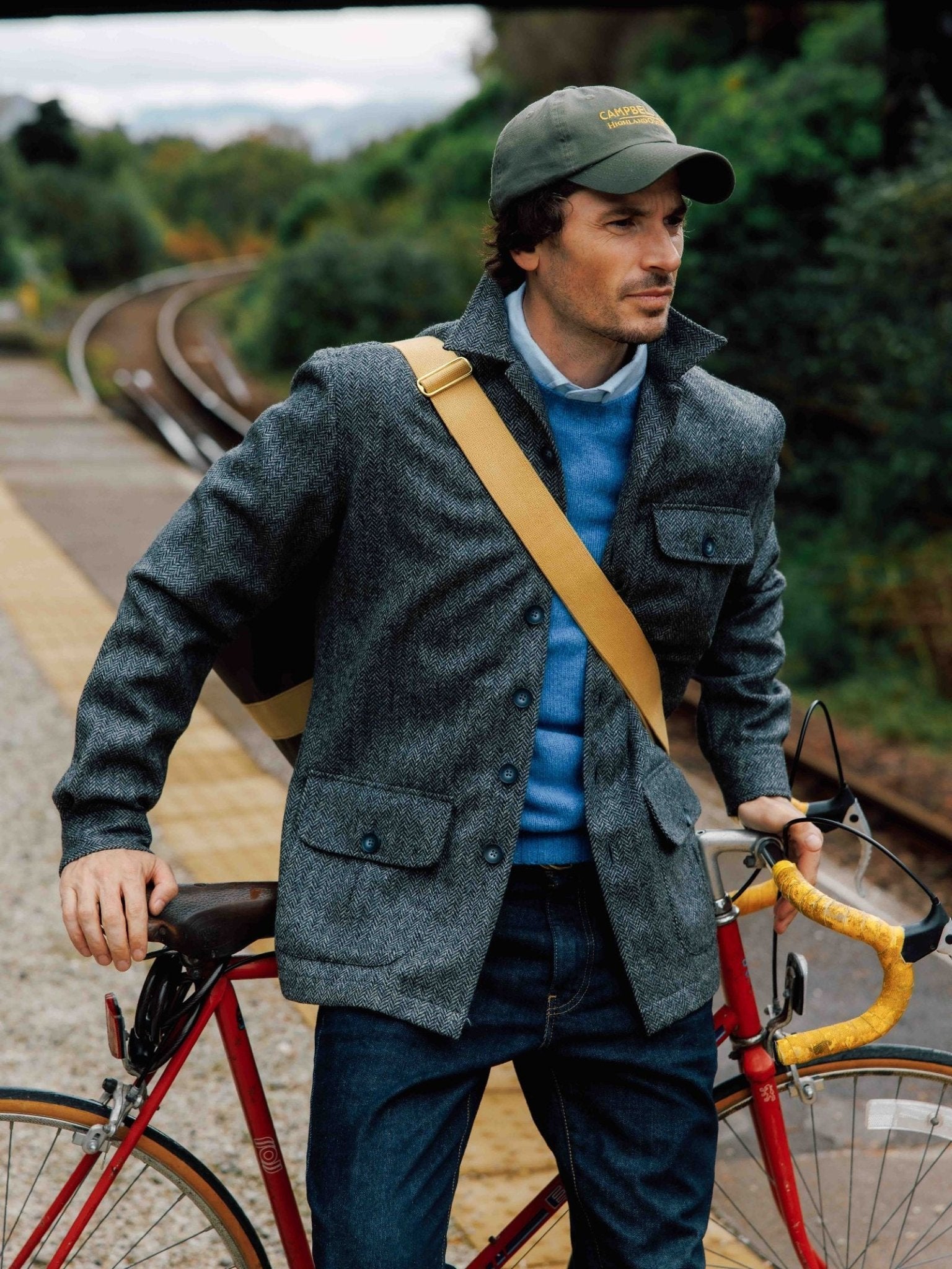 A man in the Campbell’s of Beauly Casual Tweed Shacket stands on a train platform beside a red bicycle, holding the handlebars. He pairs relaxed tailoring with a blue sweater, green cap, tan shoulder bag, and trees and tracks in the background.
