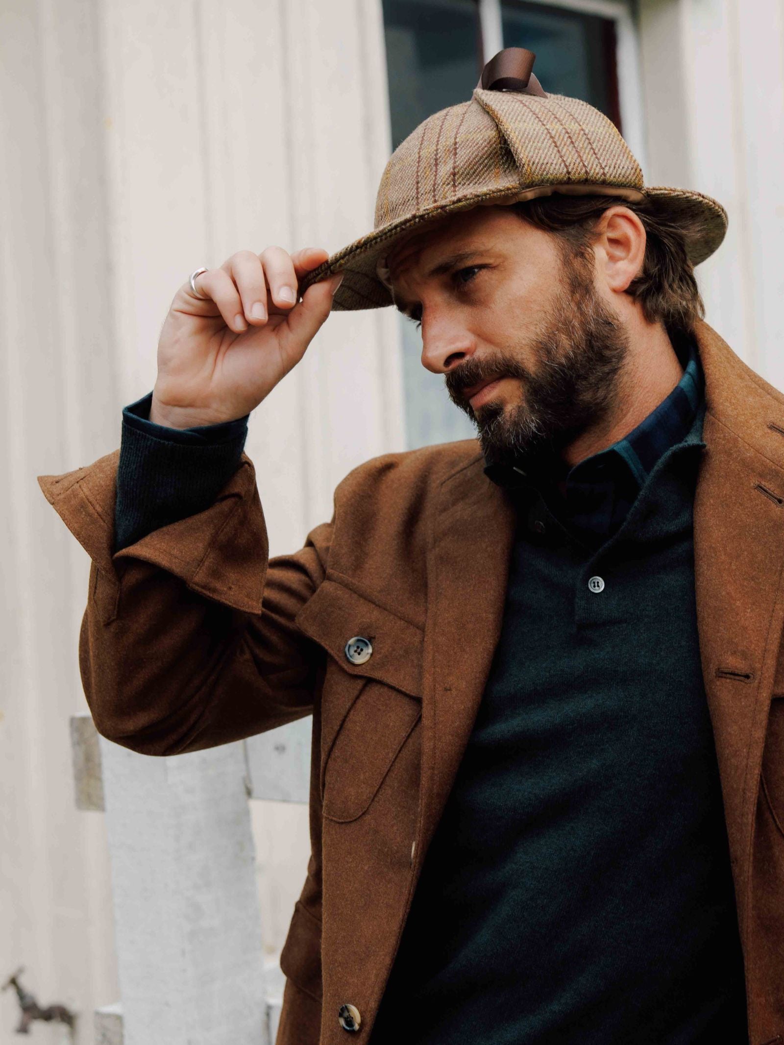 A bearded man in a brown coat tips his Campbell's of Beauly Sherlock Holmes Hat while glancing aside, standing in front of a light-colored building.
