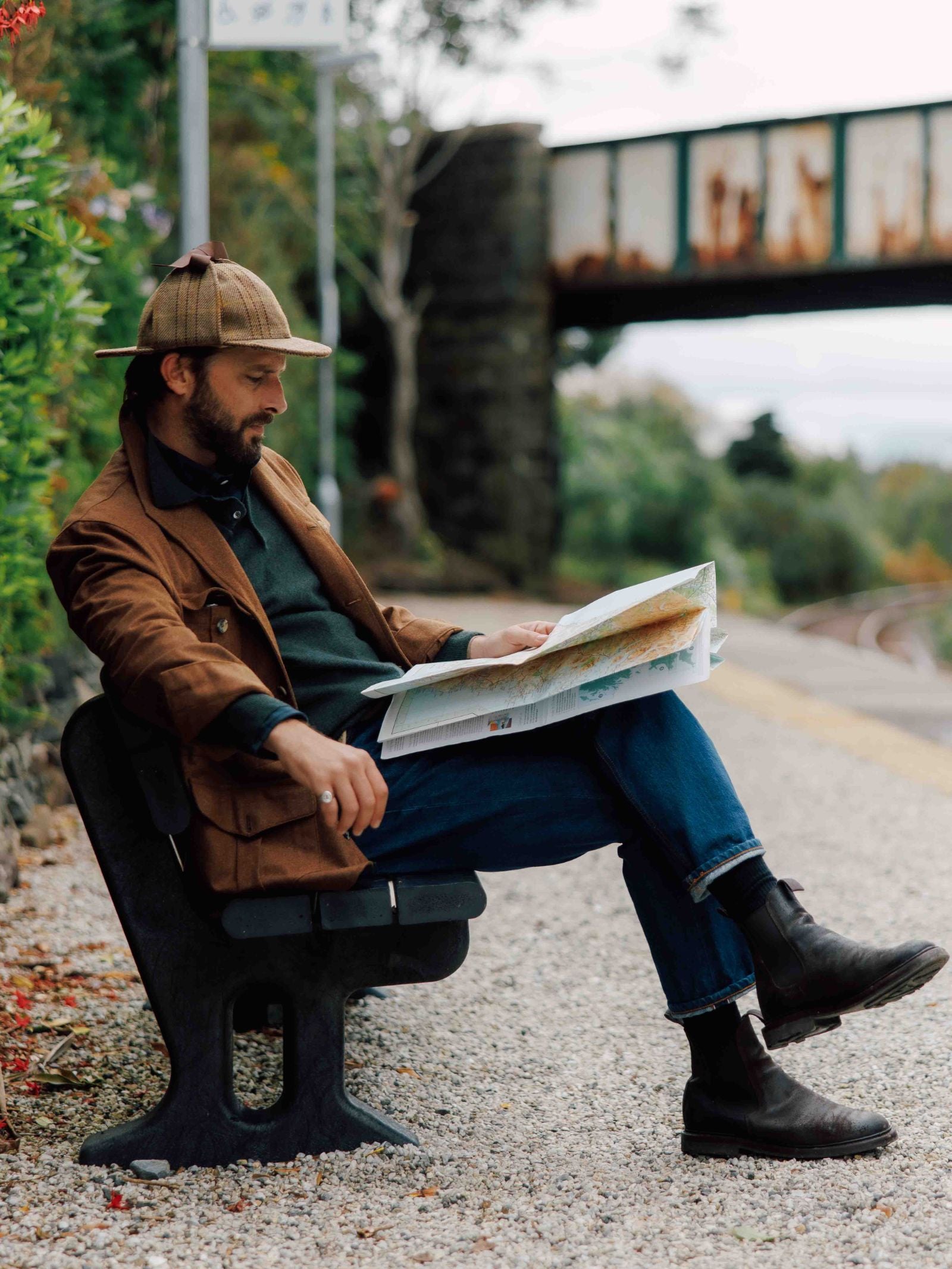 A man in a Campbell's of Beauly Field Teba Jacket, jeans, boots, and a deerstalker hat sits on a bench by railway tracks, reading a map. Greenery and an old bridge form the outdoor background.