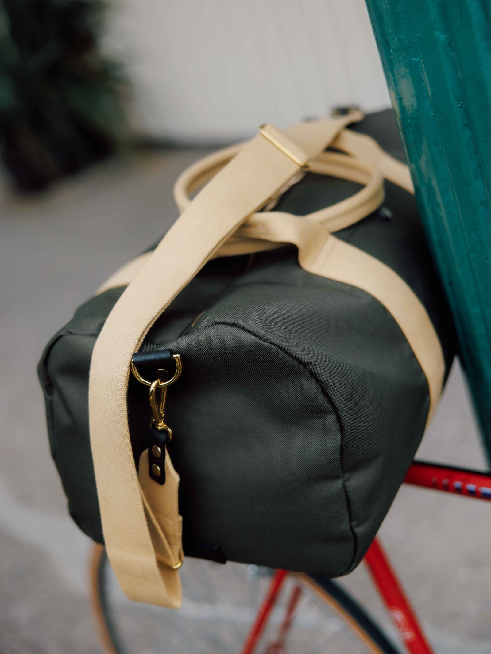 The Campbell’s of Beauly Utility Medium Duffle in dark green with beige straps is attached to the back of a red bicycle, which is propped against a green pole outdoors.