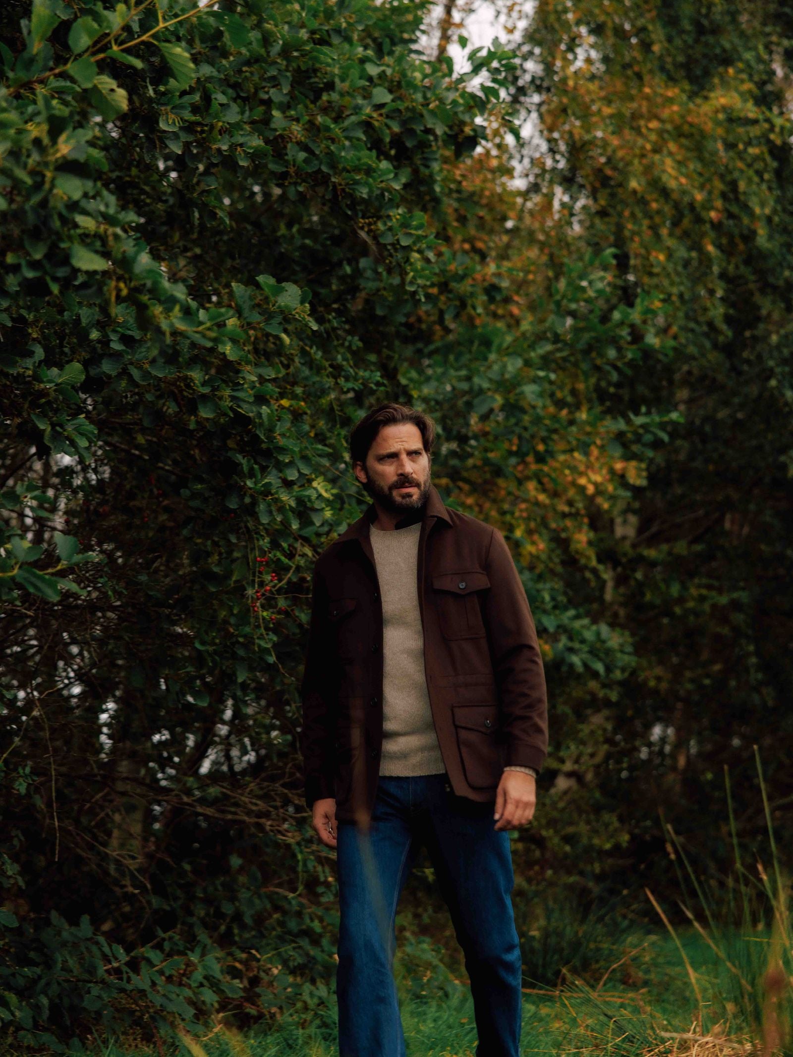A man with dark hair and a beard walks outdoors near lush greenery, wearing Campbell's of Beauly's Winter Aigas Field Jacket over a light sweater and blue jeans on a naturally overcast day.