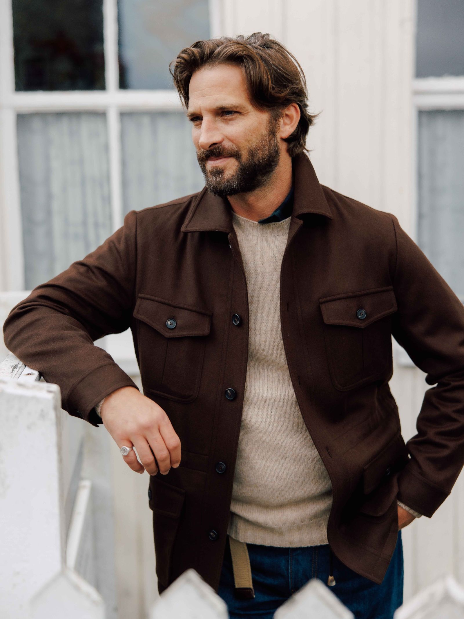 A man with brown hair and a beard stands outdoors by a white fence, wearing the Campbell's of Beauly Auld Stock Winter Aigas Field Jacket over a beige sweater and blue jeans, with windows and curtains in the background.