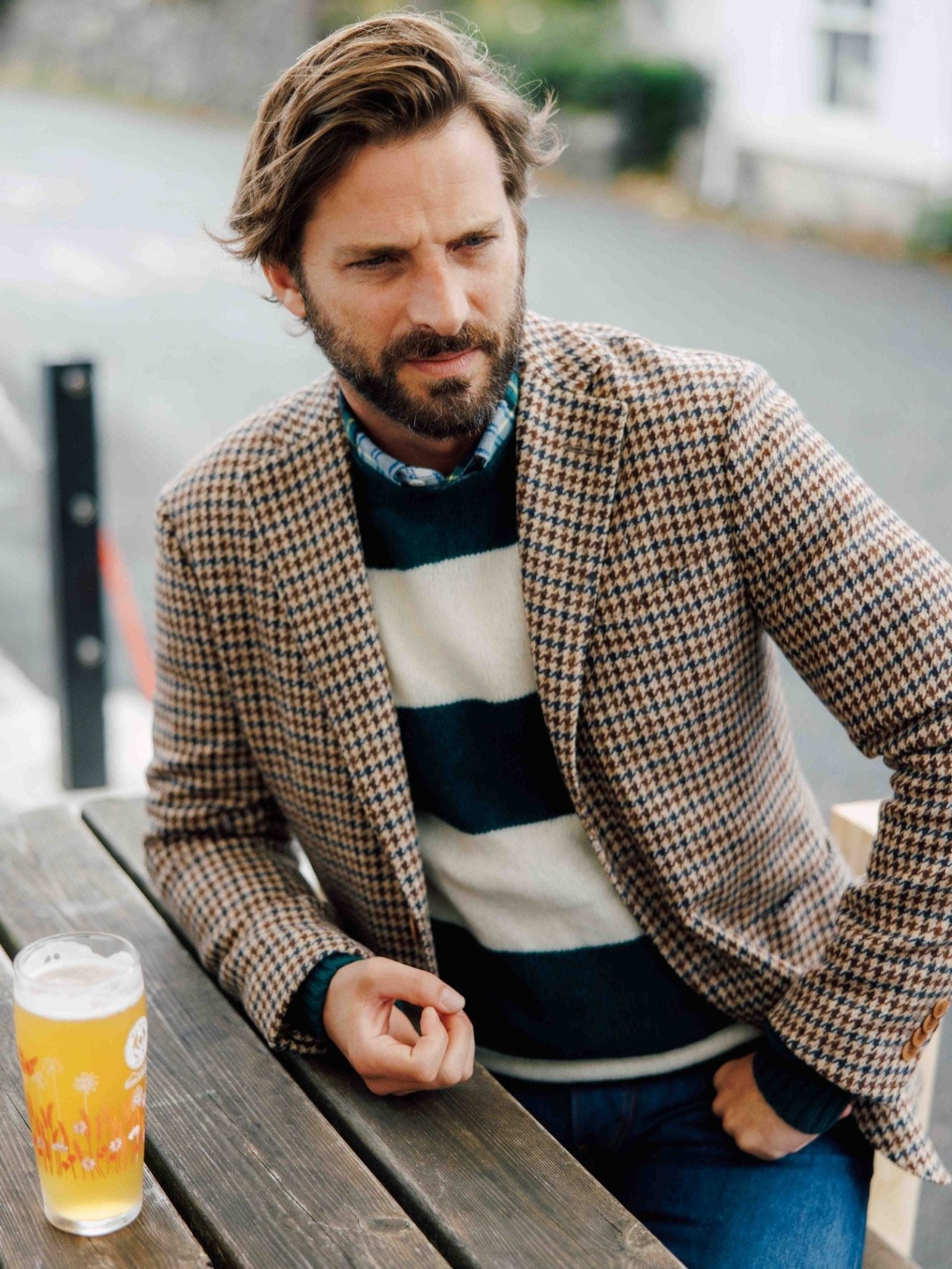 A bearded man in a brown houndstooth blazer and Campbell's of Beauly Shetland Stripe Crew Jumper sits at an outdoor wooden table with a glass of beer, gazing pensively to the side.