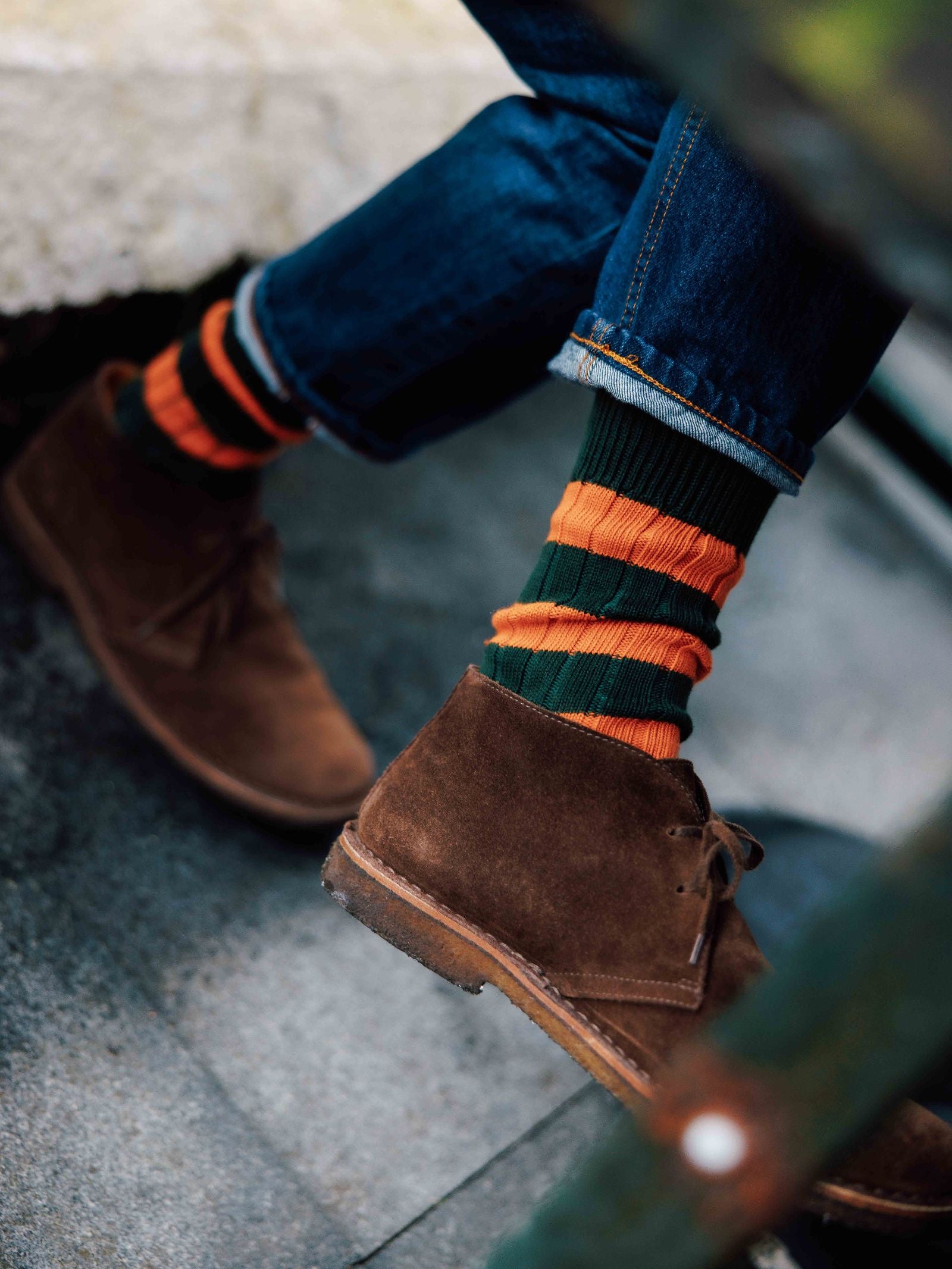 A person sits outdoors with one leg crossed, wearing cuffed blue jeans, brown suede desert boots, and Campbell's of Beauly Stripe Socks featuring green and orange stripes plus a reinforced heel and toe.