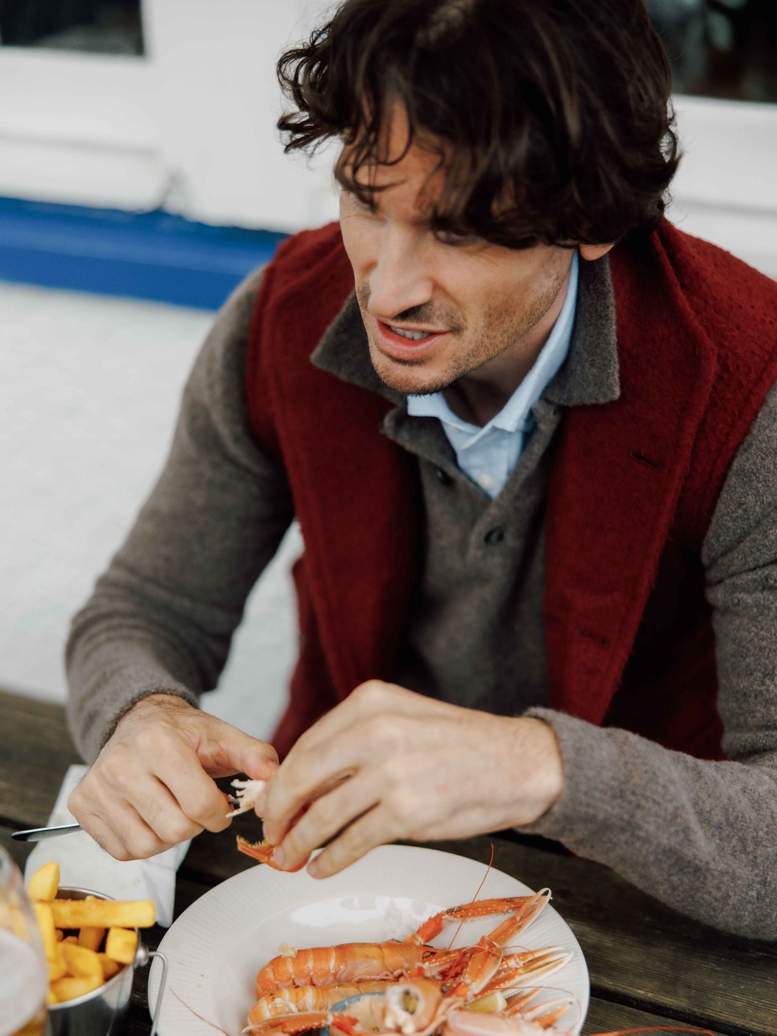 A man in a red vest over a Campbell's of Beauly Bouclé Polo Sweater sits at a table, focused as he peels shrimp from a plate, with a bowl of fries beside him.