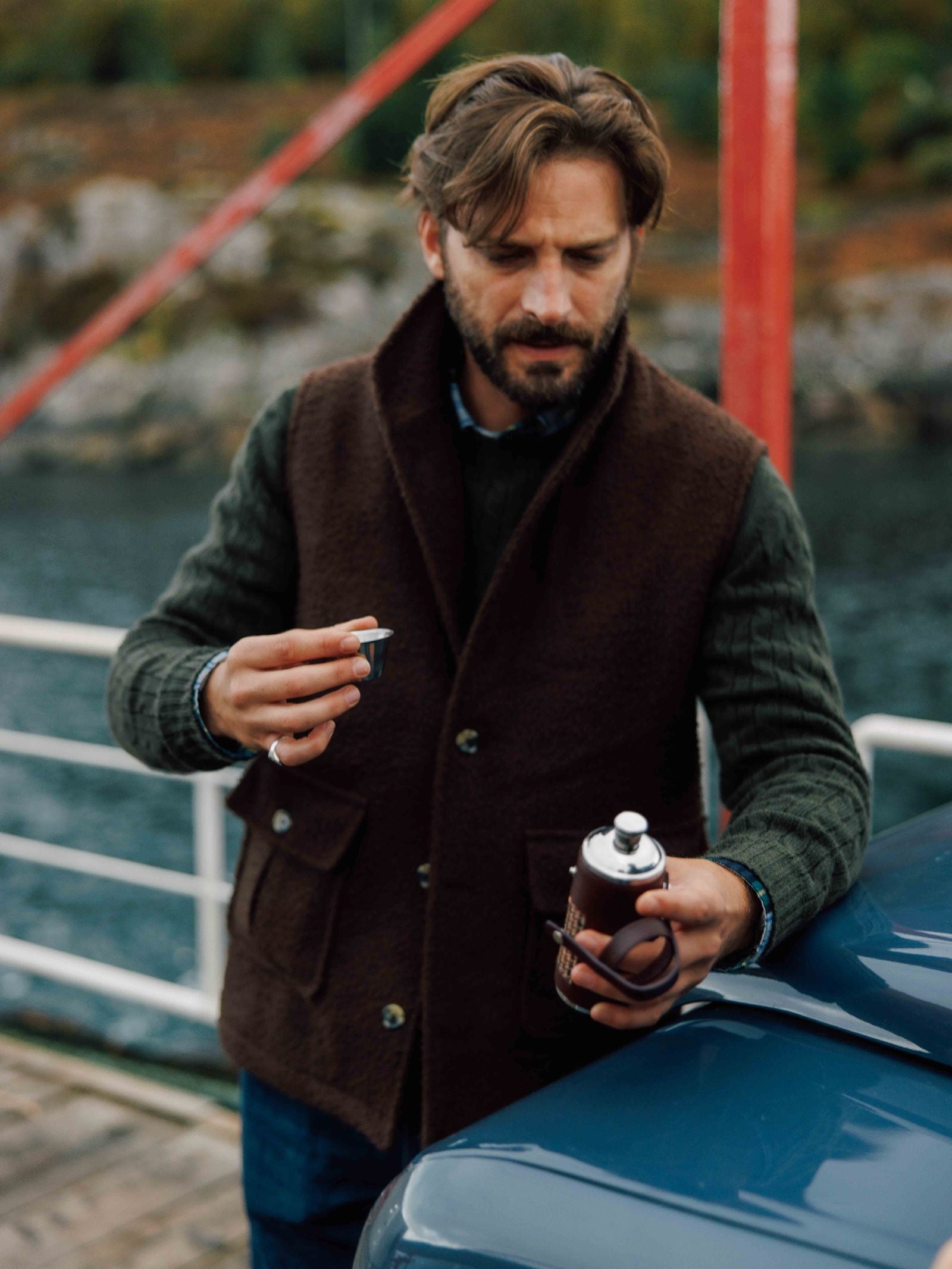 A man with brown hair and a beard, wearing a Campbell's of Beauly Casentino Fleece Gilet over a green sweater, stands by a blue car pouring a drink from a metal flask, with water and trees in the background.