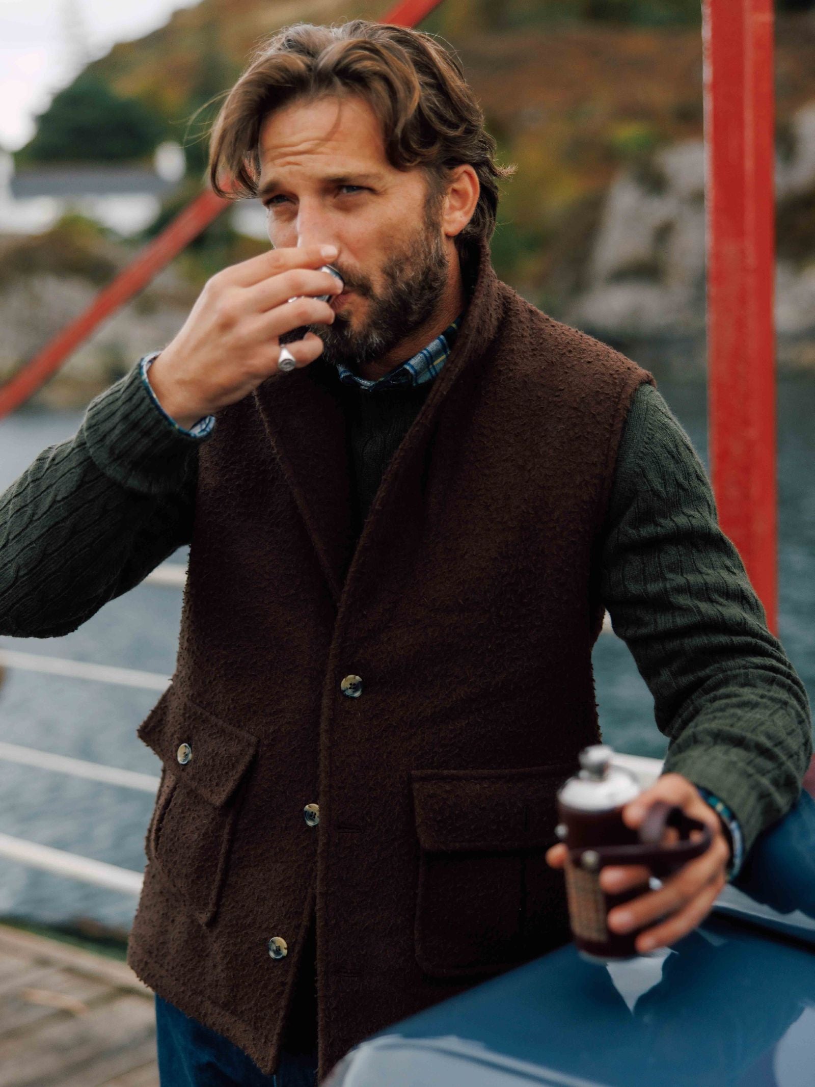 A man with wavy hair stands outdoors by the water, wearing a Campbell's of Beauly Casentino Fleece Gilet over a green sweater. He sips from a small cup and holds a metal flask, with a blurred natural background behind him.