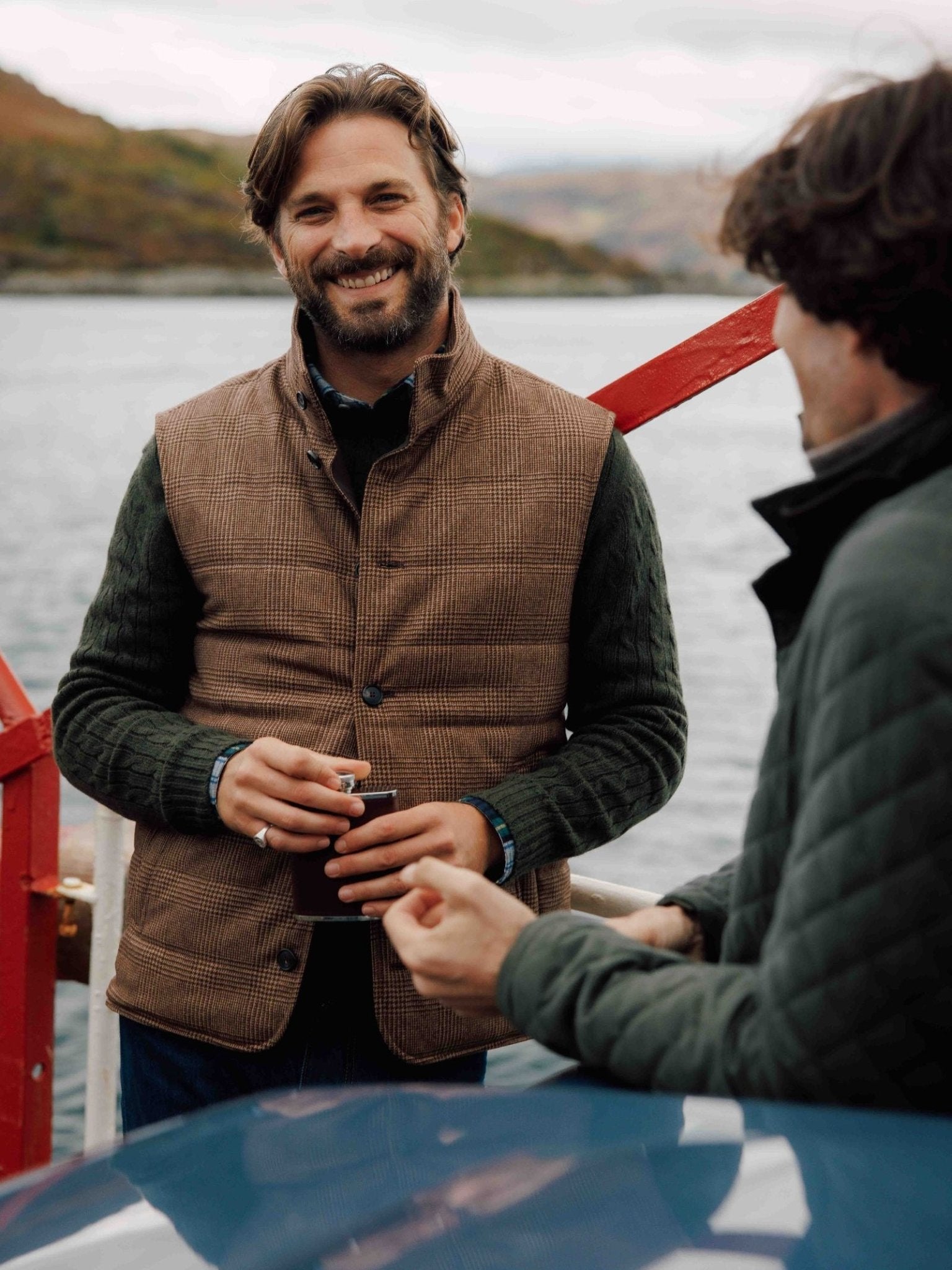 Two men stand by the water, smiling and talking. One wears the Campbell's of Beauly Cashmere Gun Club Quilted Gilet and holds a small cup; the other faces him. A red railing and hilly landscape evoke a classic country scene.