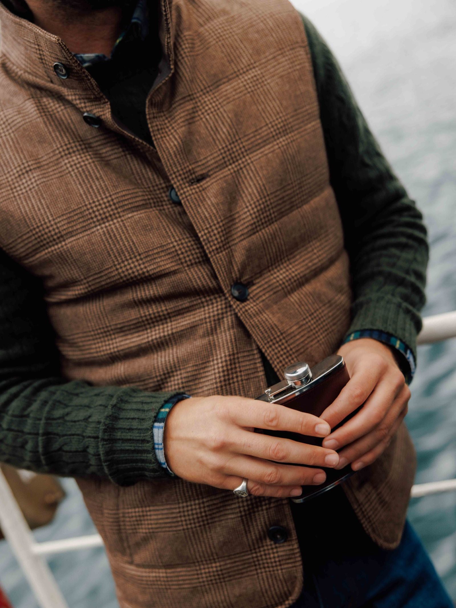 Wearing the Cashmere Gun Club Quilted Gilet by Campbell's of Beauly and a green sweater, a person holds a metal flask on a boat—an iconic country wardrobe look inspired by classic British style.