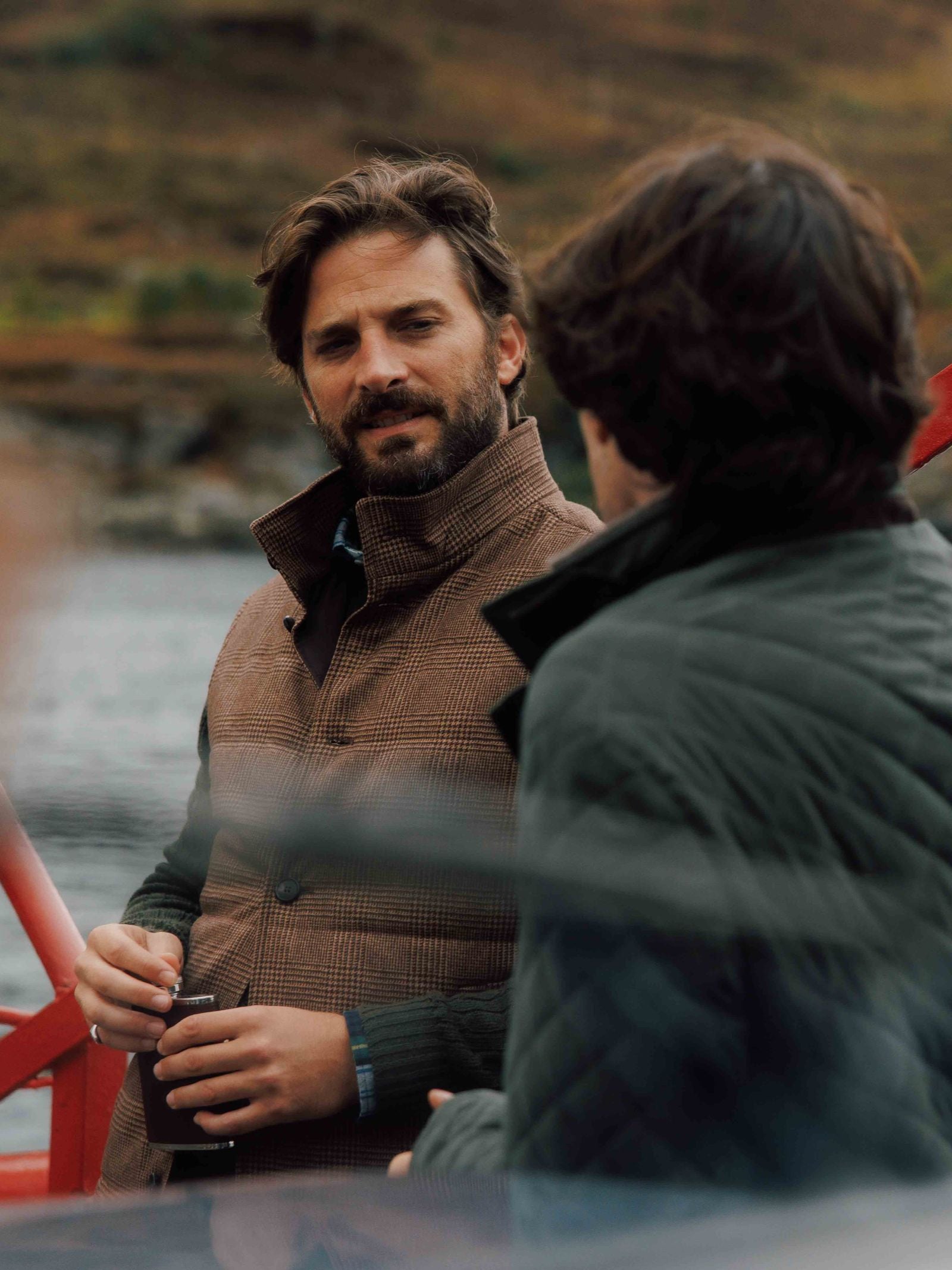 Two men chat outdoors by the water. One holds a drink can, wearing a brown jacket with British tweed details; the other, seen from behind, wears a Campbell's of Beauly Cashmere Gun Club Quilted Gilet. Blurred natural scenery is in the background.