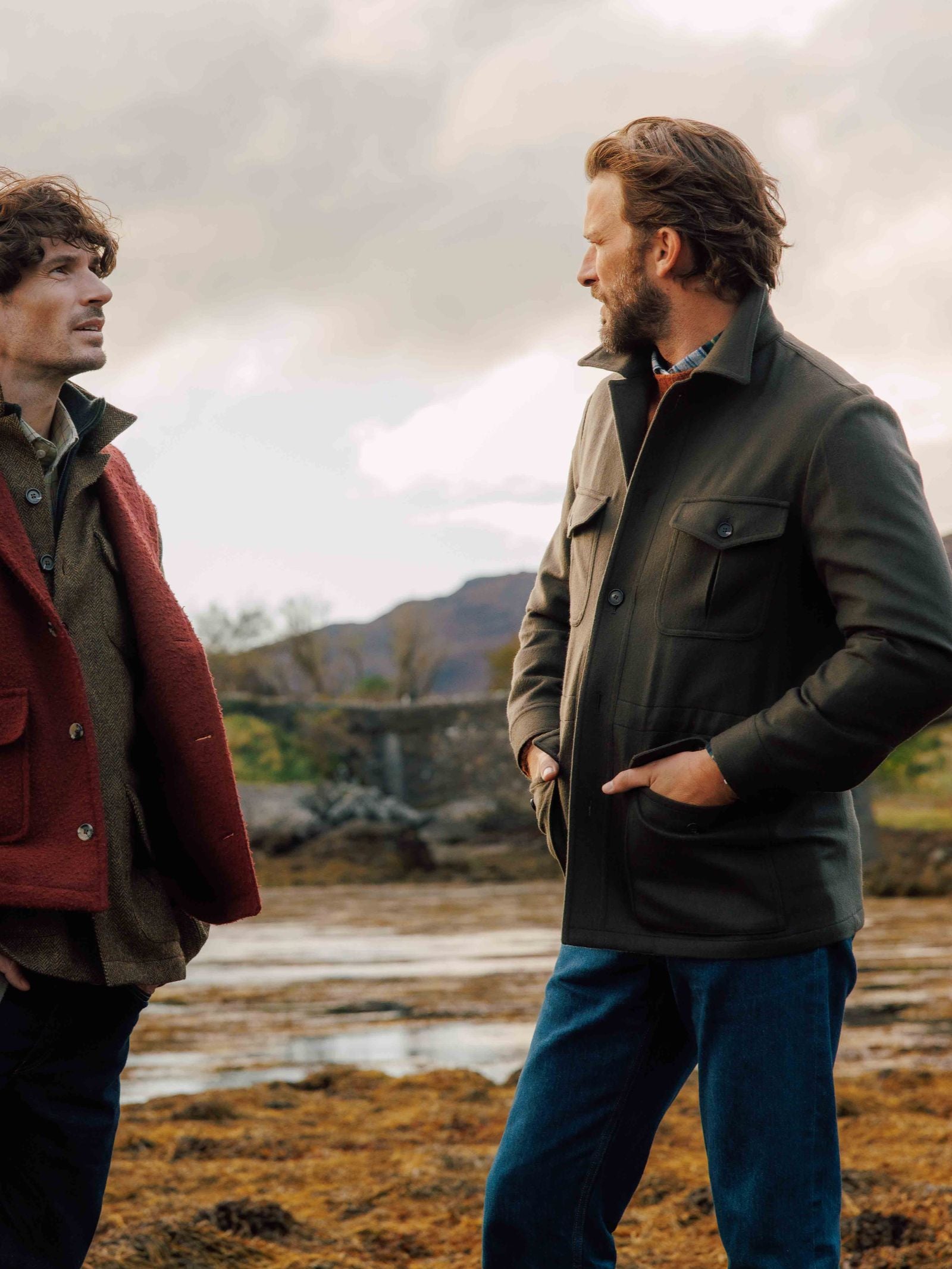 Two men stand and talk near a stone bridge with hills and a cloudy sky, both wearing Campbell's of Beauly Winter Aigas Field Jackets—one in red, one in green—hands in pockets, in a scenic rural setting.