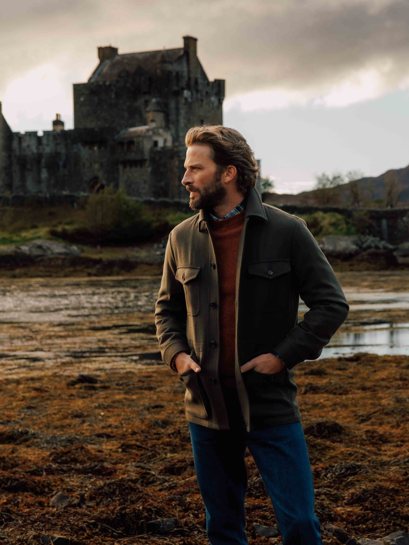 Standing on rocky ground near a historic stone castle, a bearded man looks to the side with his hands in his pockets, wearing blue jeans, a sweater, and the Campbell's of Beauly Winter Aigas Field Jacket.