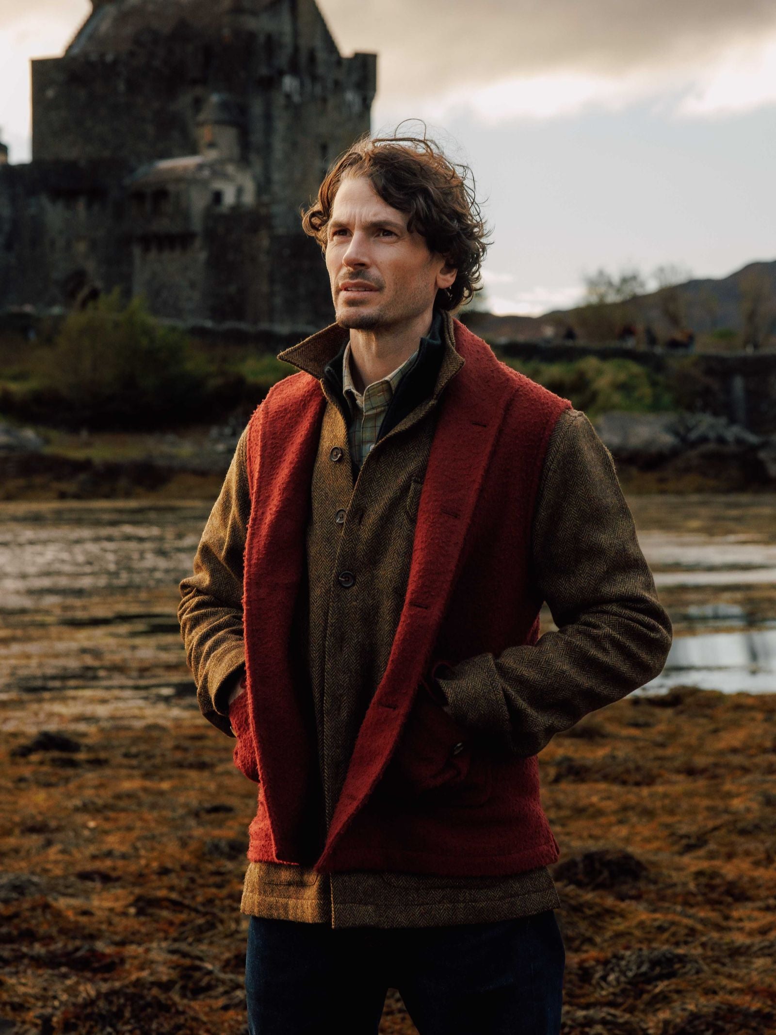 A man with wavy brown hair stands outdoors by a historic stone castle, wearing a Campbell's of Beauly Casentino Fleece Gilet over his brown jacket. His hands are in his pockets as he stands on earthy, uneven ground under soft light.