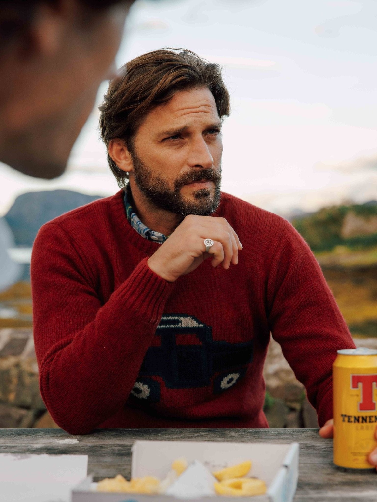 A bearded man in a Campbell's of Beauly Unisex Land Rover Defender Jumper sits at an outdoor table, holding a can of Tennent’s beer with food boxes in front, while another person is partly visible in the foreground.