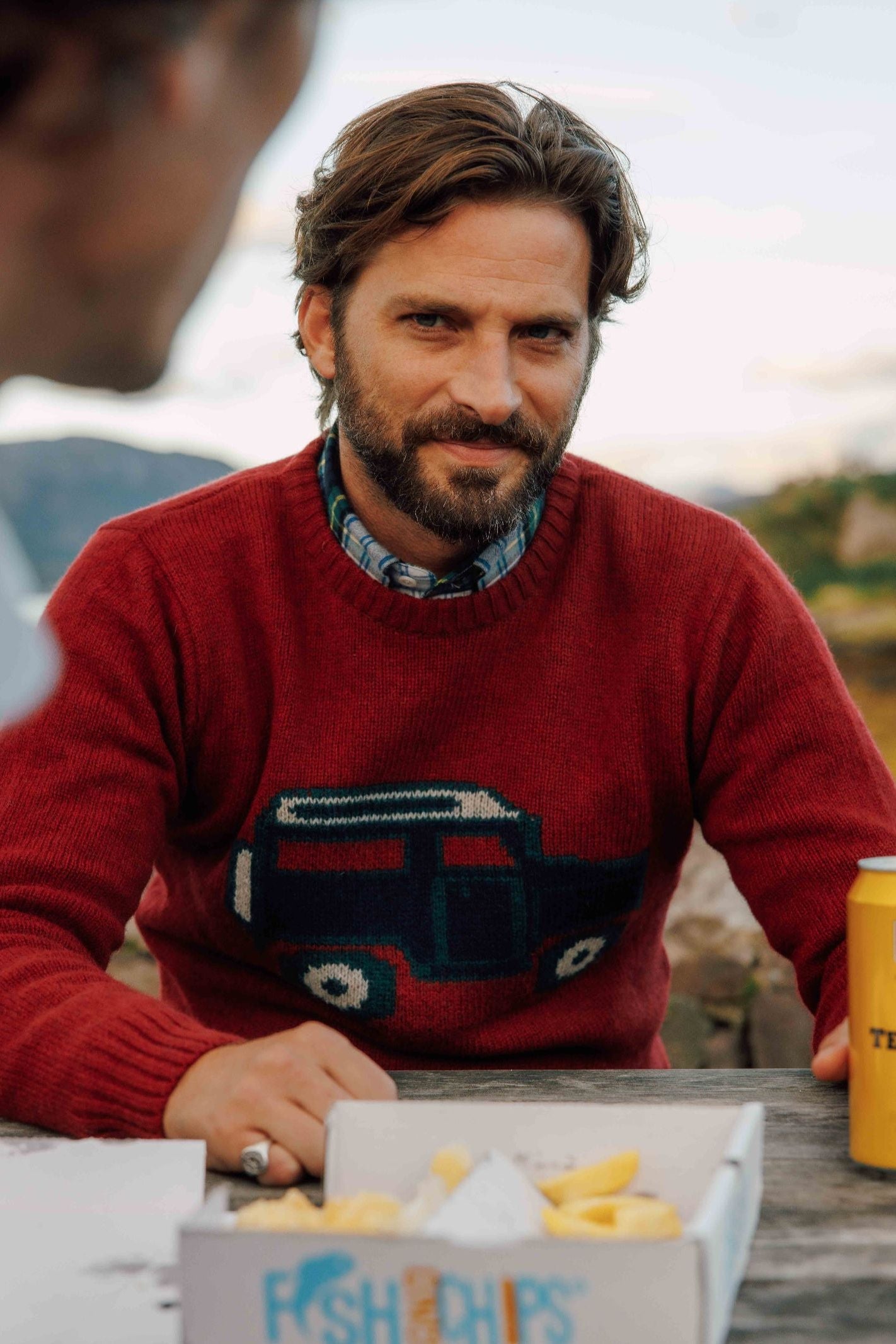 A bearded man wearing the Campbell's of Beauly Land Rover Defender Jumper sits outdoors at a picnic table with a food box of fish and chips and a yellow can of Tennent's beer in front of him, gazing off-camera.