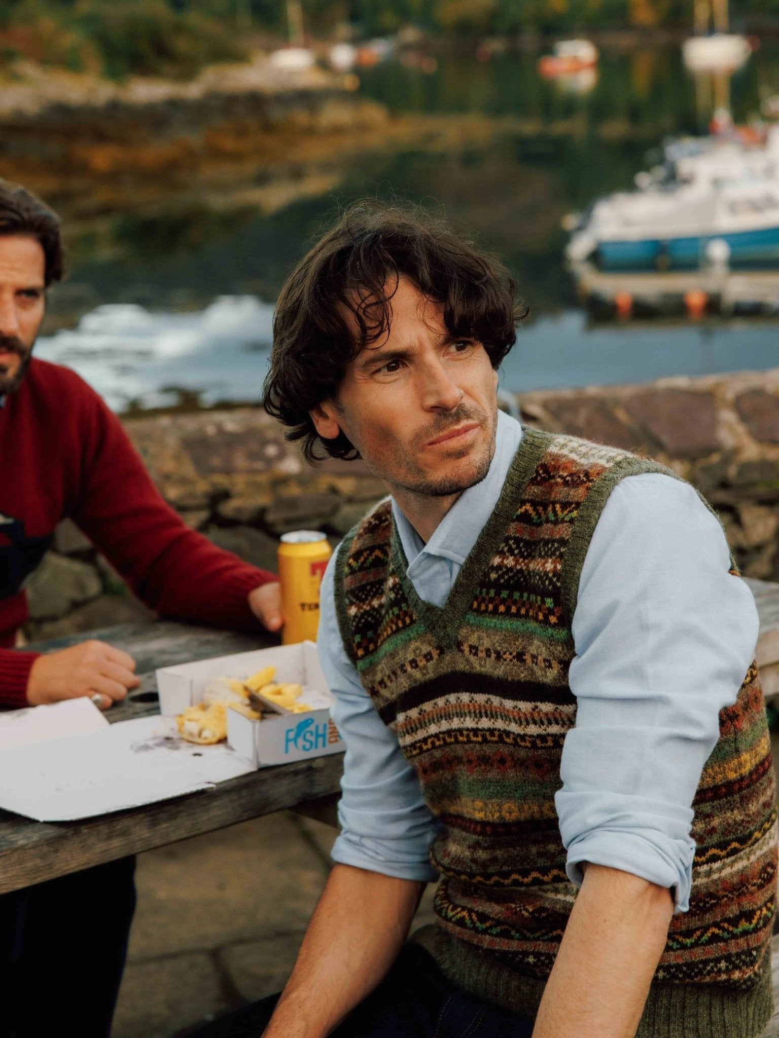 A man with wavy dark hair wears a Campbell's of Beauly Fairisle Slipover over a light blue shirt as he sits at an outdoor picnic table by the water, with food, drinks, and boats in the background.