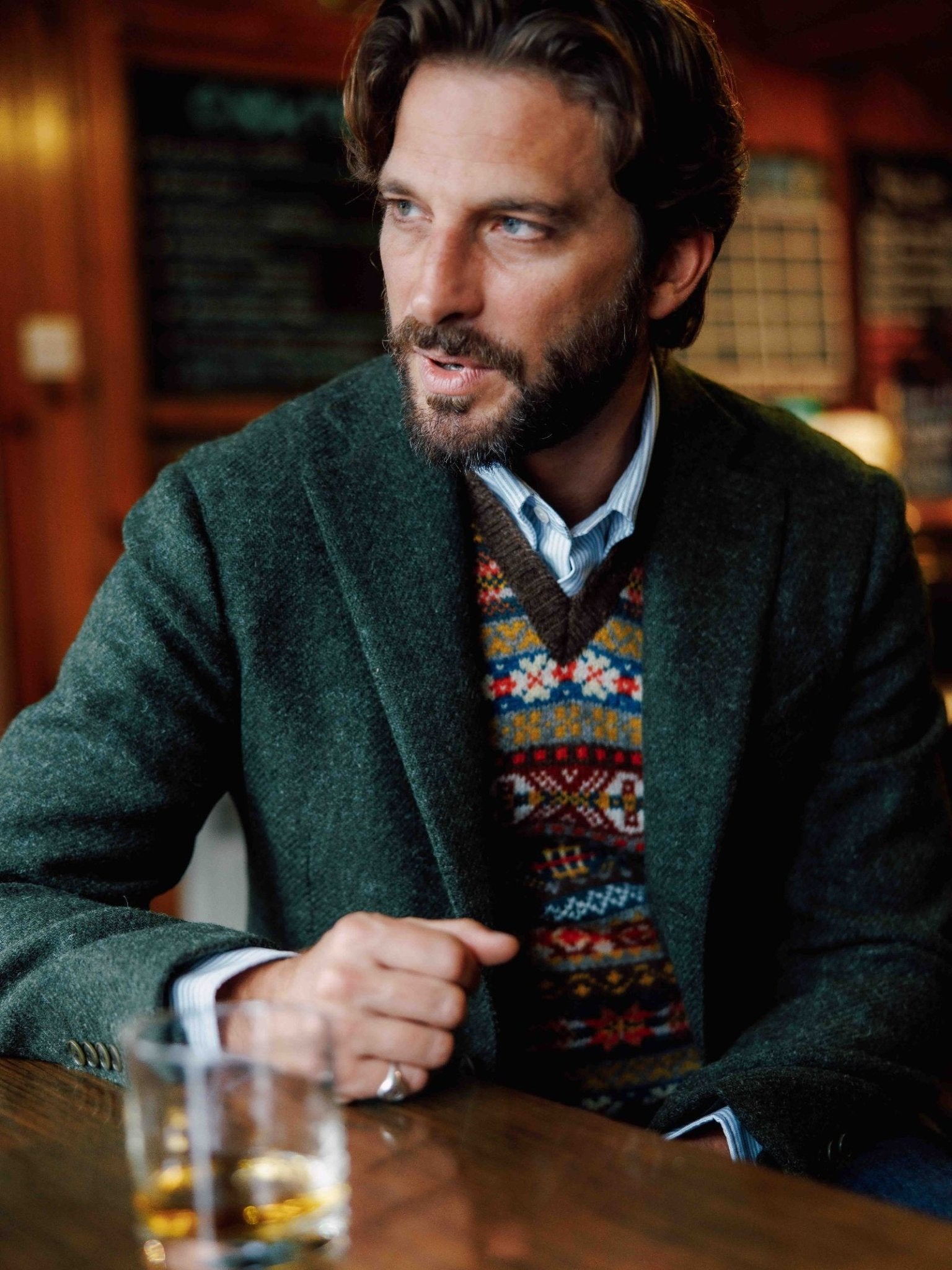 A man with medium-length brown hair and a beard wears a regular fit Campbell’s of Beauly Harris Tweed Jacket and a patterned sweater over a collared shirt, seated at a wooden table with whiskey in a cozy, warmly lit setting.