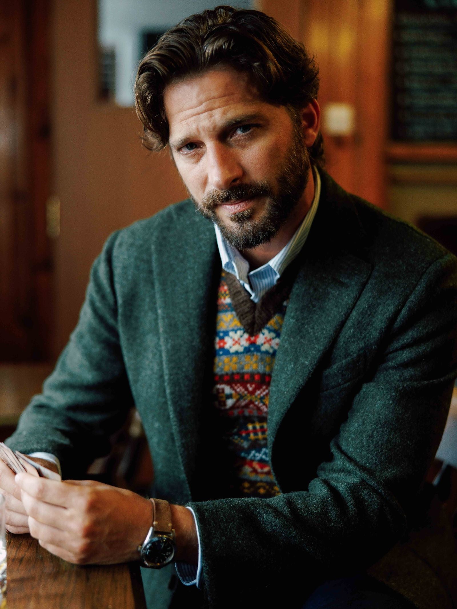 A man with wavy brown hair and a beard sits at a wooden table, thoughtfully gazing at the camera in a Campbell's of Beauly Harris Tweed Jacket layered over a colorful sweater vest and blue shirt.