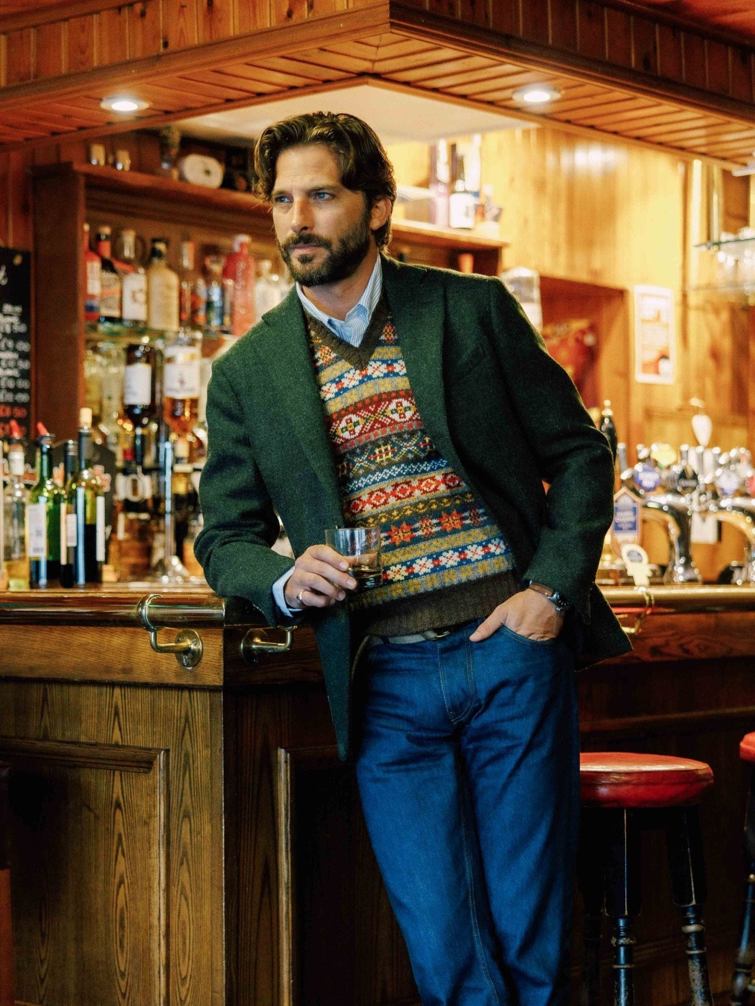 A man with brown hair and a beard stands at a wooden bar, holding whiskey. He wears a Campbell's of Beauly Harris Tweed Jacket in regular fit over a patterned vest, blue shirt, and jeans. The bar has bottles and wood-paneled walls.