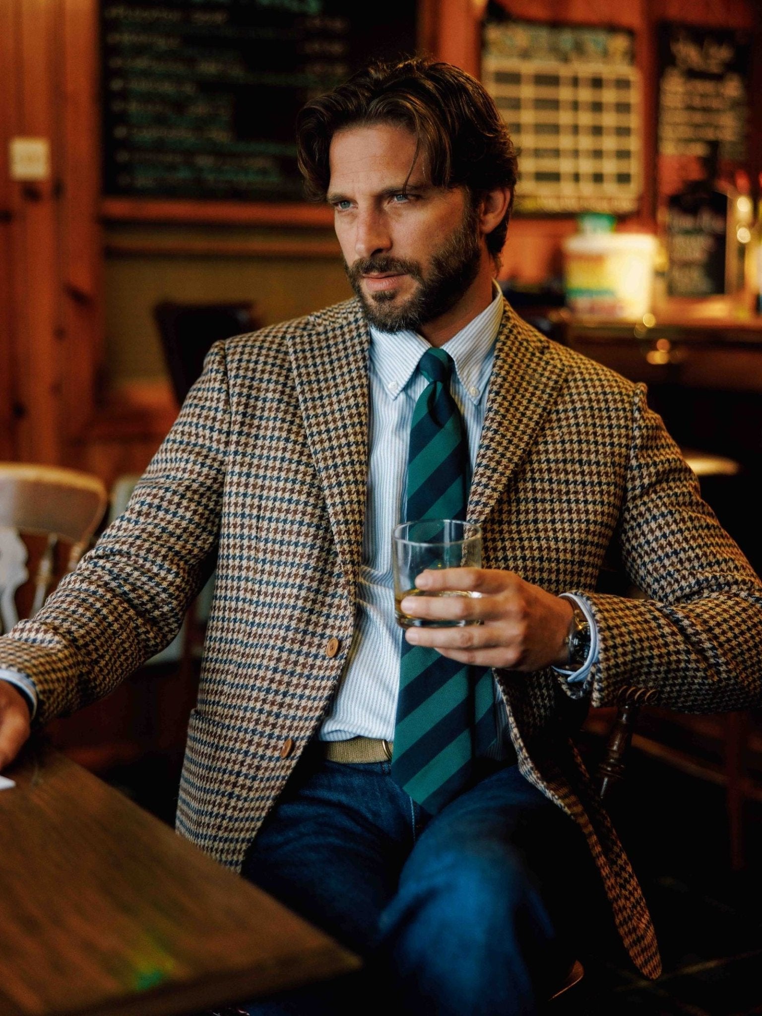 A man with dark hair and a beard sits at a wooden table in a cozy bar, wearing a houndstooth blazer, striped shirt, Campbell's of Beauly Green & Navy Stripe Tie, and jeans, holding a glass of whiskey and looking to the side.