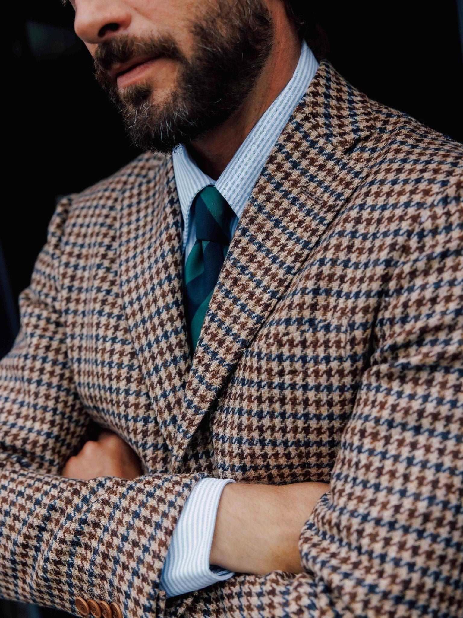 A bearded man stands with arms crossed, wearing Campbell's of Beauly's Auld Stock Oxford Shirt in light blue, paired with a brown and blue houndstooth suit and a dark green and blue striped tie.