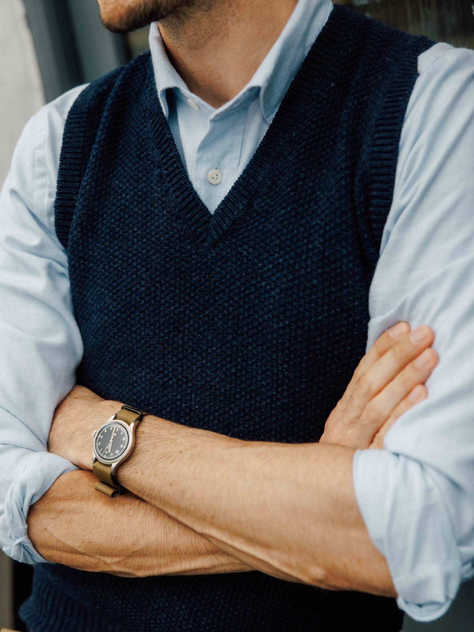 A man with folded arms wears a light blue shirt under a Campbell’s of Beauly Moss Stitch Knitted Slipover, made in the UK. He has a brown-strap watch on his left wrist, and his face is partially out of frame.