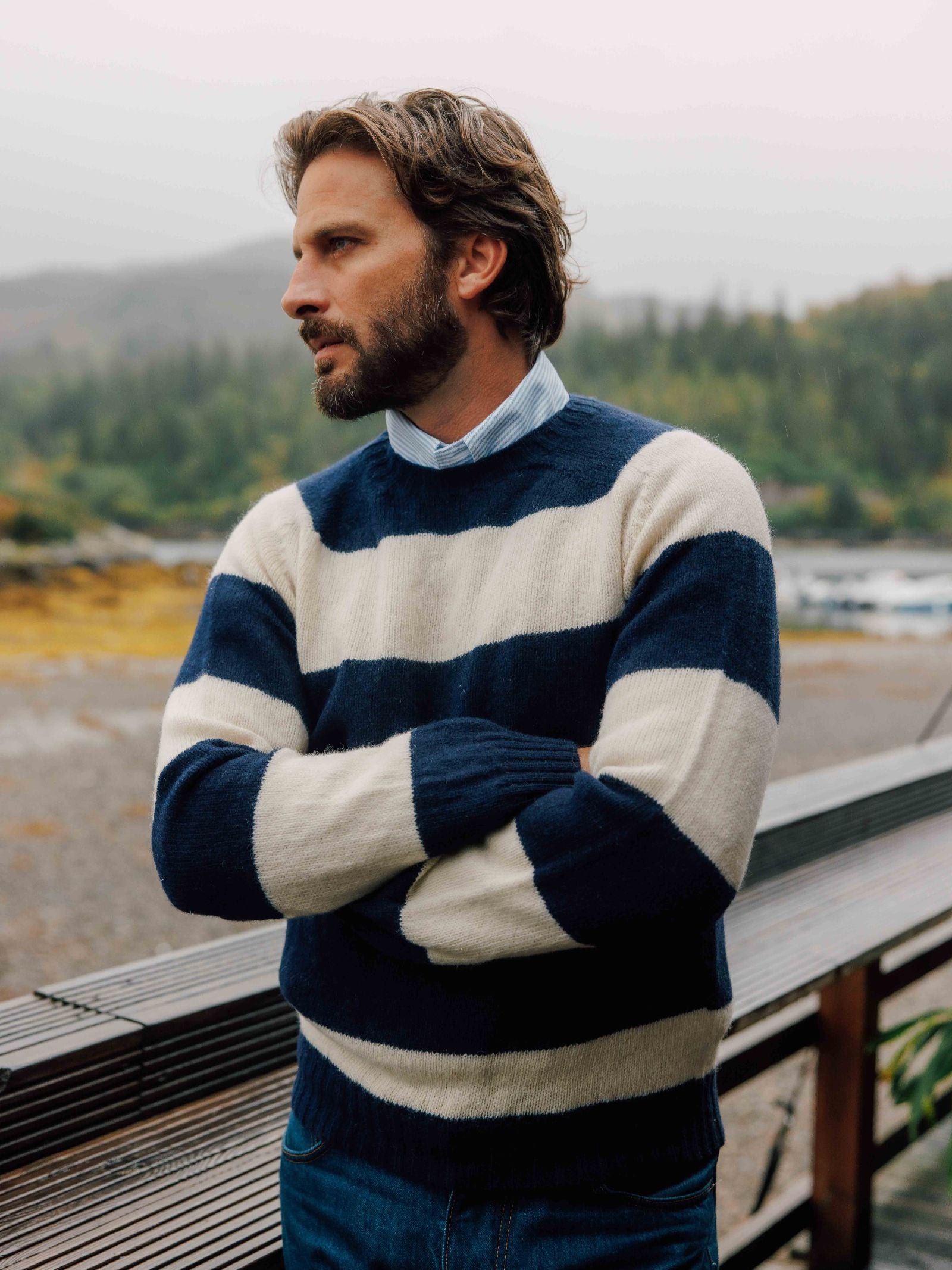 A man with a beard and medium-length hair stands outdoors with arms crossed, wearing the Shetland Stripe Crew Jumper by Campbell's of Beauly over a collared shirt. Misty trees and mountains form the background.