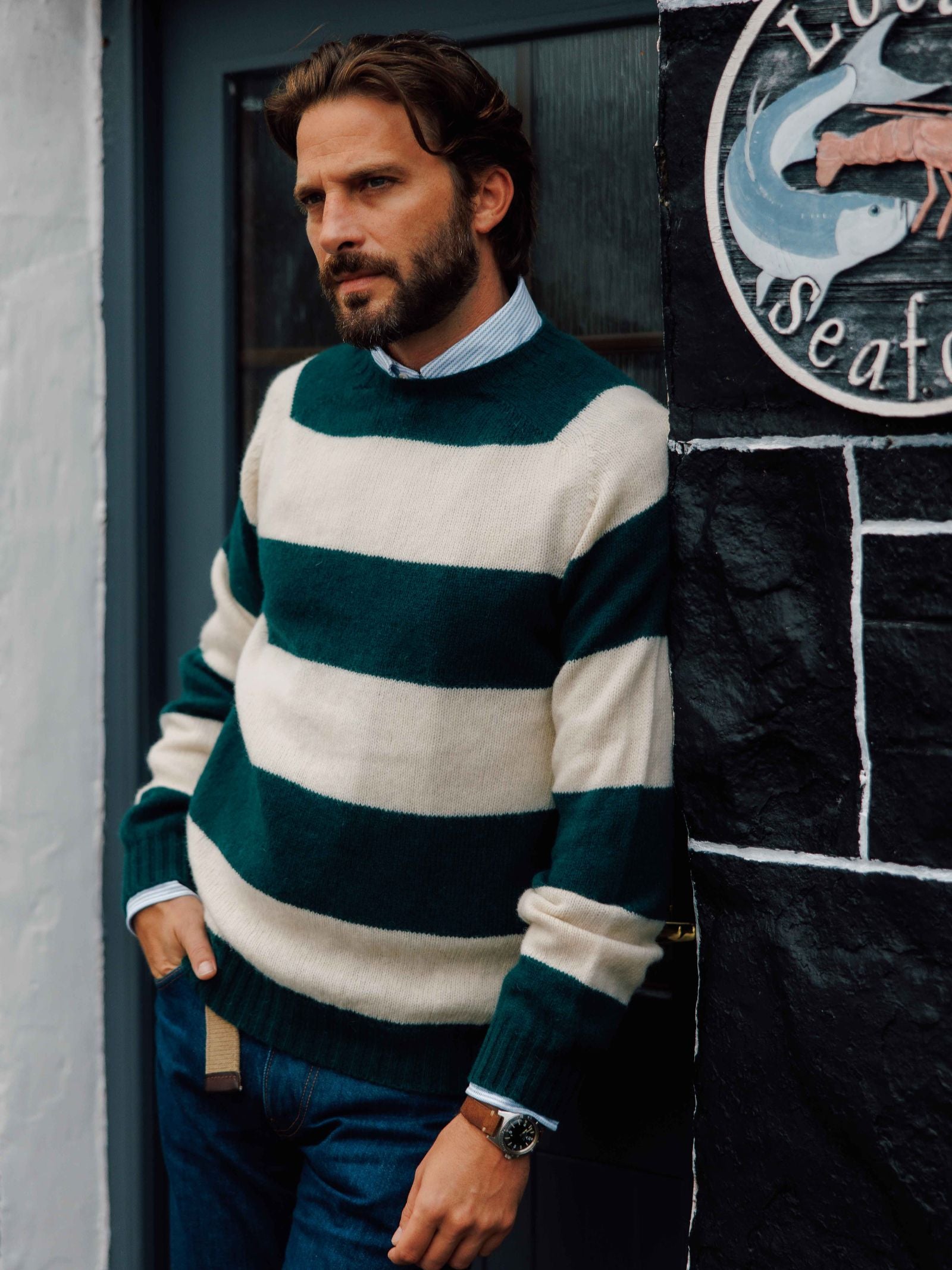 A man with brown hair and a beard wears the Campbell's of Beauly Shetland Stripe Crew Jumper over a collared shirt and jeans, leaning against a black and white wall near a door, gazing thoughtfully into the distance.