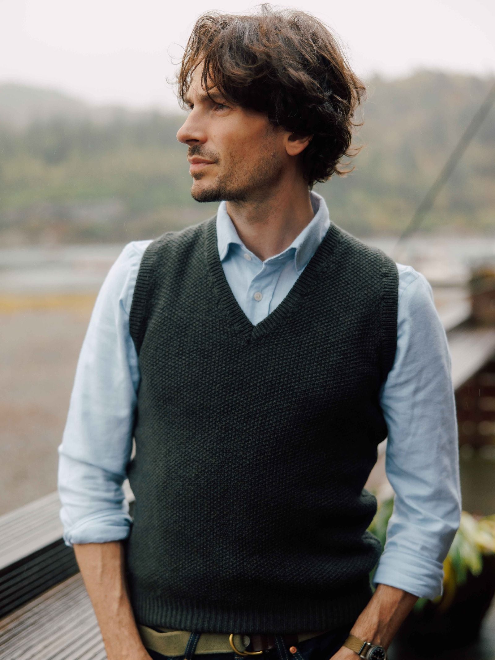 A man with wavy brown hair and a light blue shirt wears the Campbell's of Beauly Moss Stitch Knitted Slipover, made in the UK, while standing outdoors. A misty, natural landscape is blurred in the background as he looks to the side.